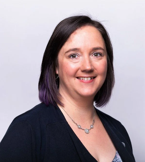 Professional woman with shoulder-length dark hair and purple tips, smiling, wearing a black top and a silver necklace, against a plain light background.