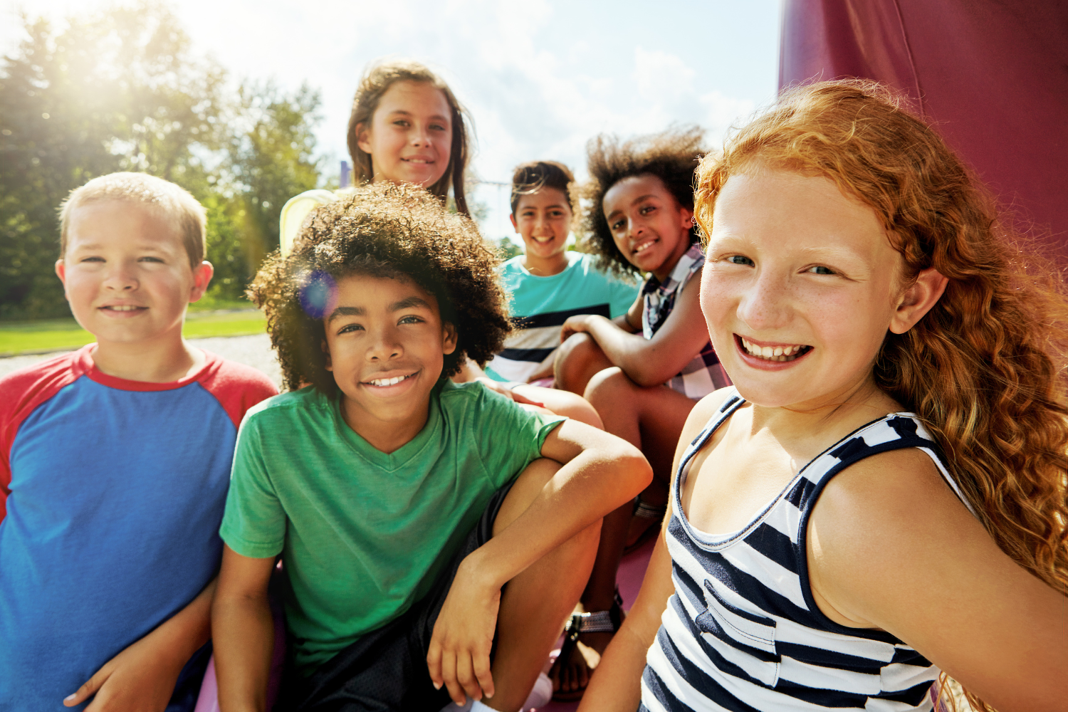 Group of six smiling children outdoors on a sunny day.