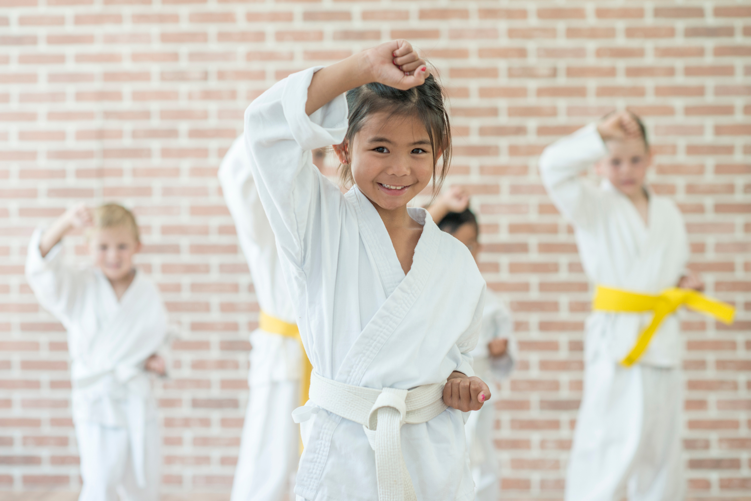 Young girl in a karate uniform with a yellow belt raising her fist in a karate training class with other children, all in karate uniforms, against a brick wall background.
