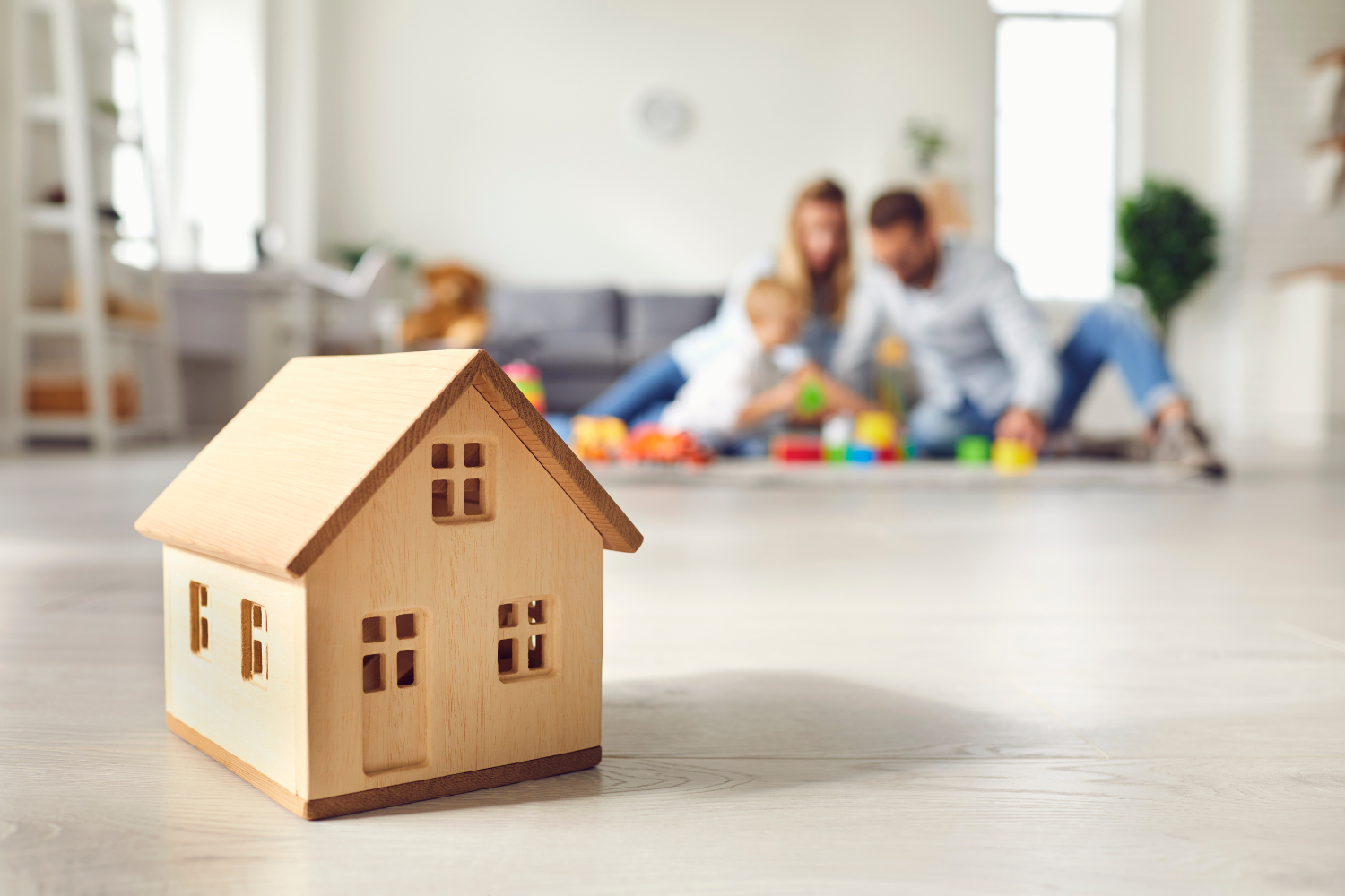 A small wooden house model on a light-colored floor with a family of three playing with toys in the background.