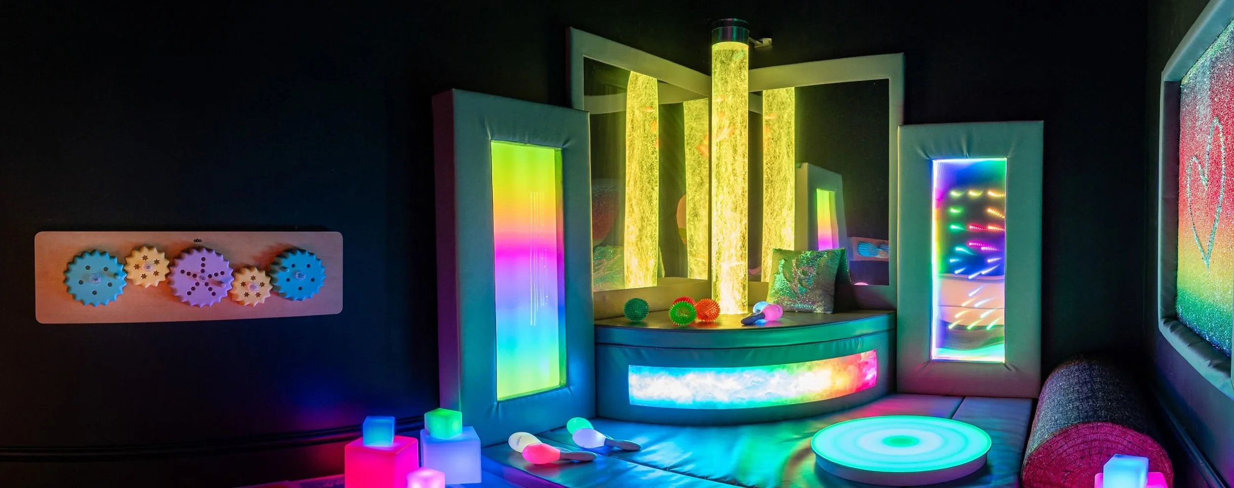 A colorful, illuminated play area with neon lights, textured panels, and soft seating, featuring vibrant light fixtures and playful decorations.