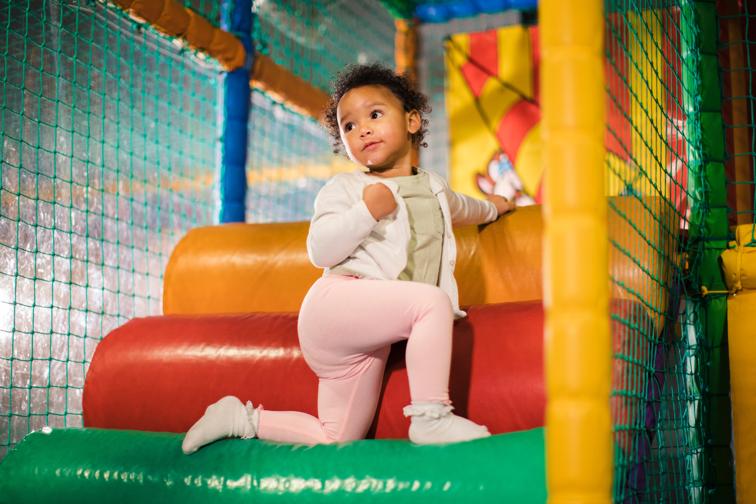 A young girl with curly hair, dressed in a light green shirt, white cardigan, and pink pants, is climbing on an indoor play structure with colorful padded surfaces and netting.