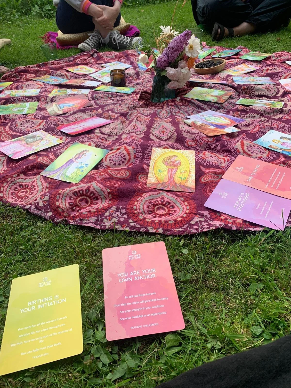 A colorful outdoor gathering setup with a patterned cloth spread on grass, tarot or oracle cards with spiritual illustrations, and motivational cards with messages about personal growth and self-awareness. There are flowers in a vase and some bowls on the cloth, and a few people sitting around it.