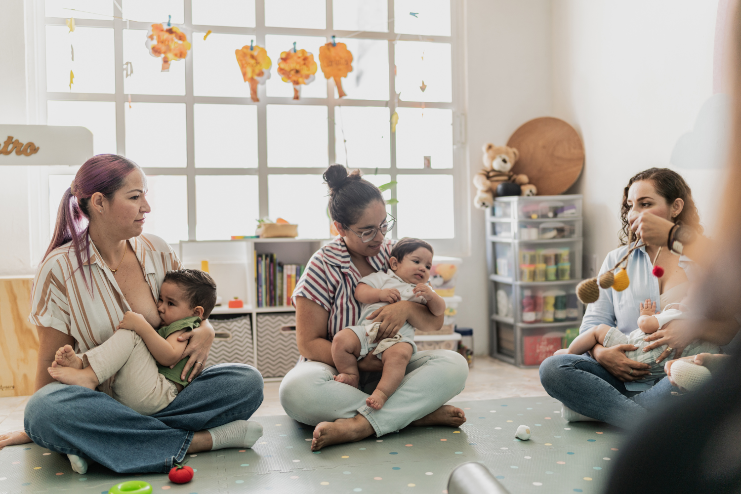 Three women sitting on the floor with babies, playing and interacting in a brightly lit room with decorations and storage shelves.