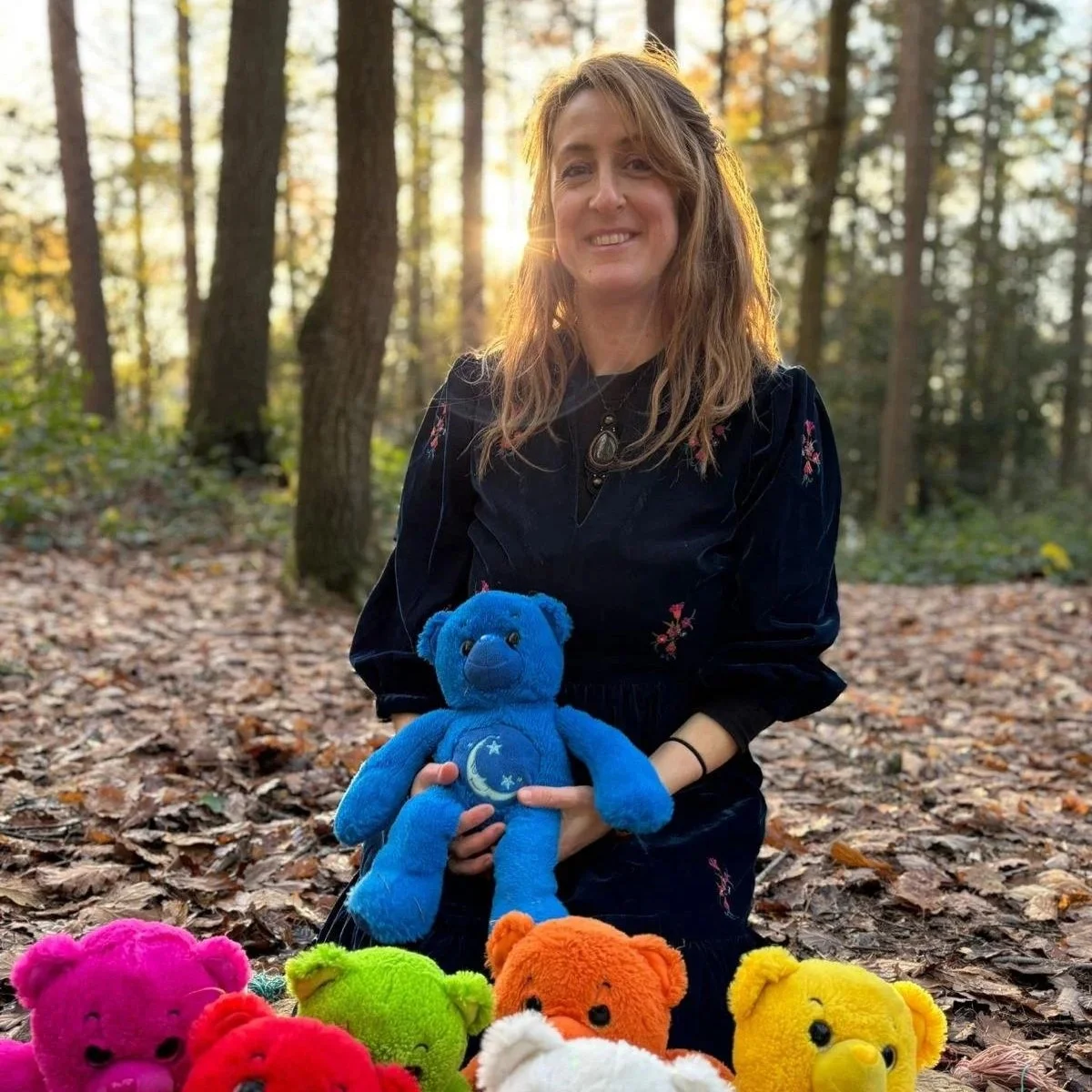 A woman with long wavy hair sitting in a wooded area during fall, holding a blue teddy bear with a moon and star design on its belly, surrounded by colorful teddy bears on the ground.