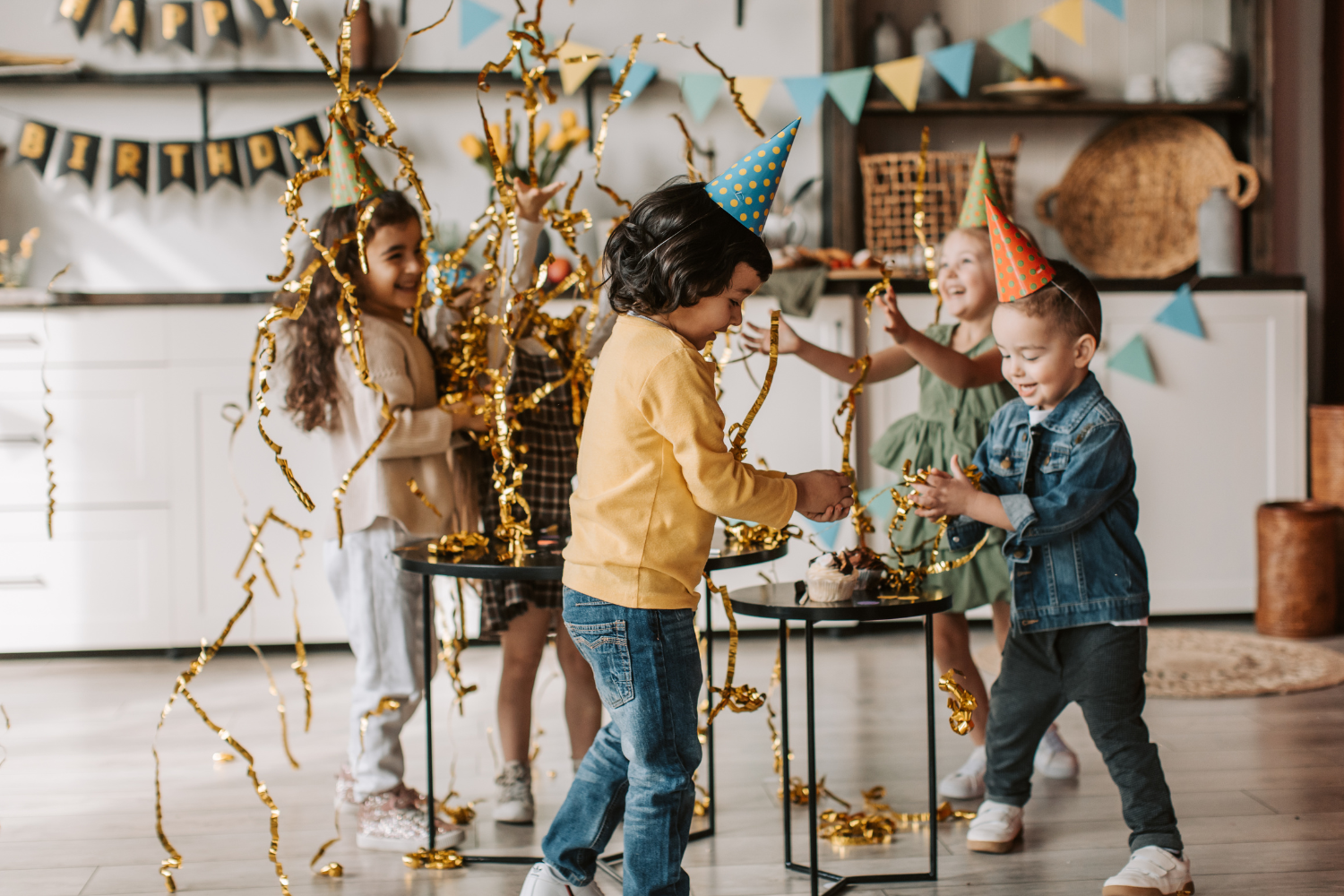 Children celebrating birthday party with party hats, confetti, and a cake in a decorated living room.