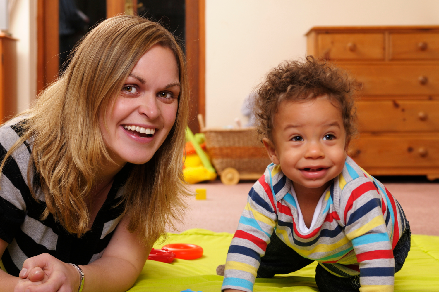 A smiling woman and a toddler with curly hair playing on the floor in a bedroom.