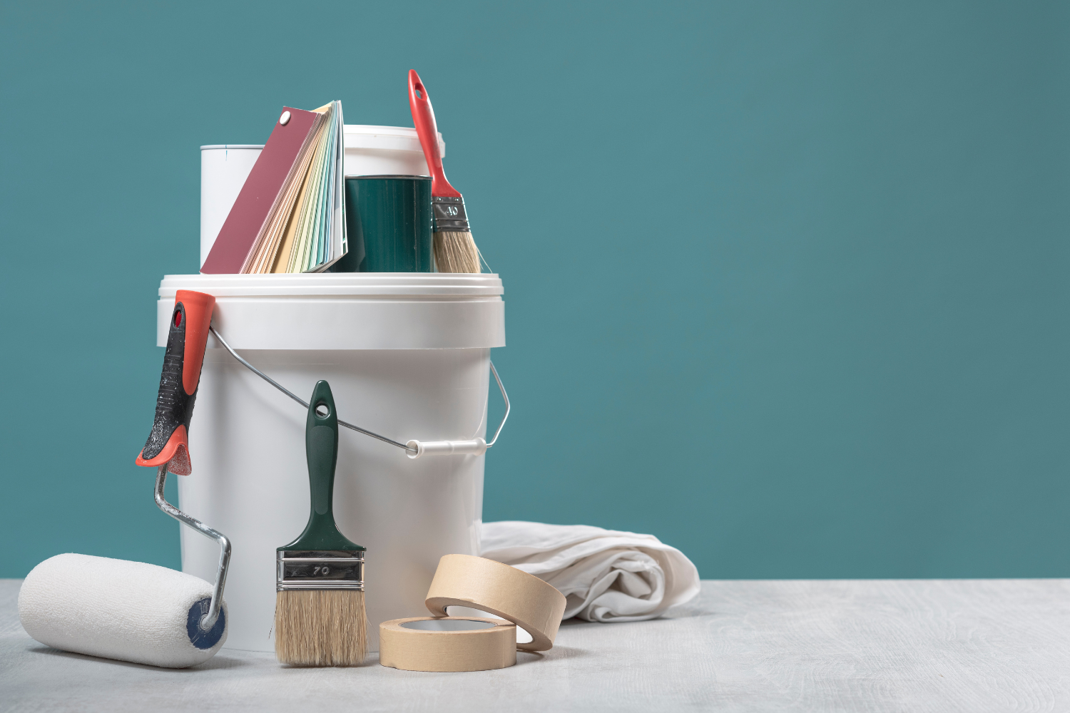 Painting supplies including a white paint bucket, paint rollers, paintbrushes, painter's tape, and a cloth on a gray surface against a teal wall