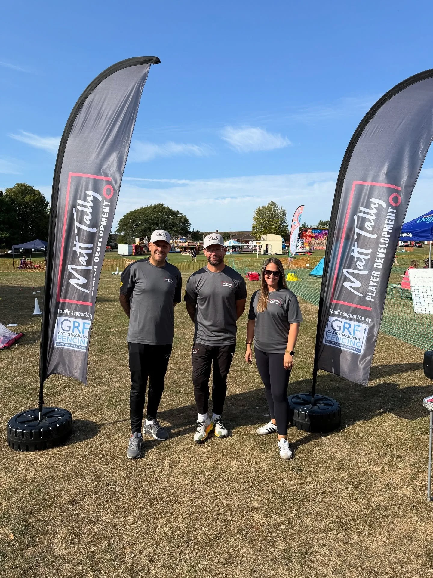 Three people standing outdoors in front of two tall banners that read "Matt Talbot" and "Player Development." They are smiling and wearing athletic clothing, with a field, tents, and other event structures visible in the background under a blue sky.