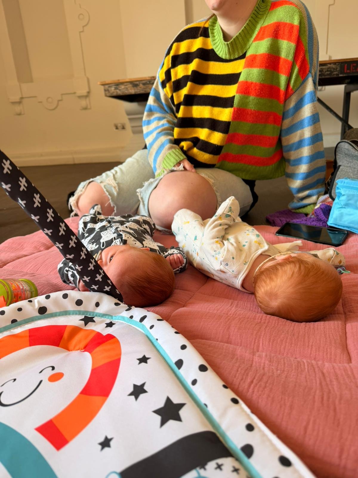 Two babies with red hair lying on a pink blanket on the floor, with one adult woman in a colorful striped sweater nearby. The babies are on their backs, looking around, with baby toys and a phone nearby.
