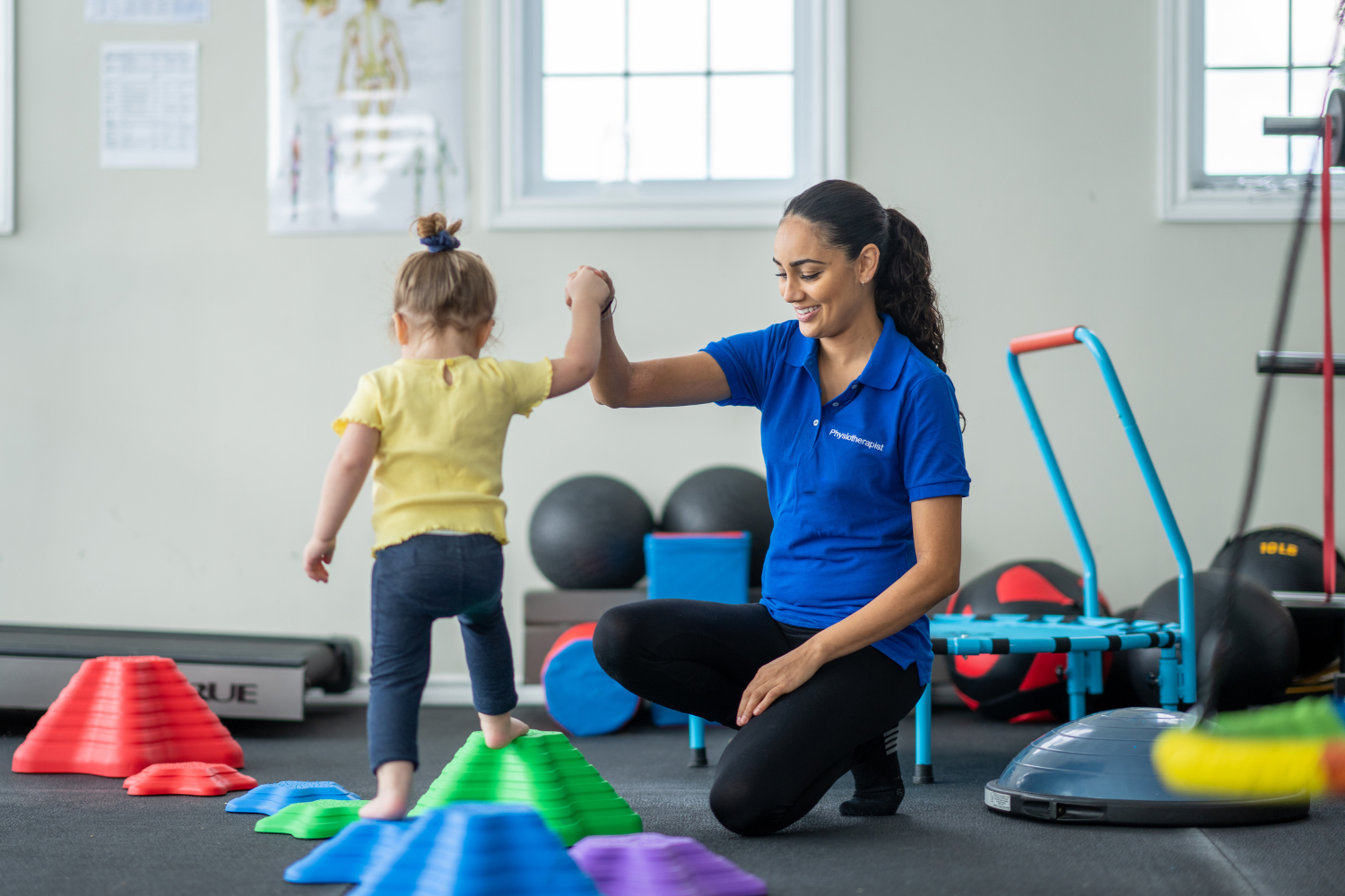 A female physical therapist helping a young girl walk on colorful balance cones in a therapy gym with fitness equipment in the background.