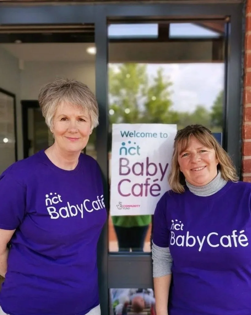 Two women wearing purple 'NCT Baby Cafe' shirts standing outside a building with a sign that says 'Welcome to NCT Baby Café' behind them.