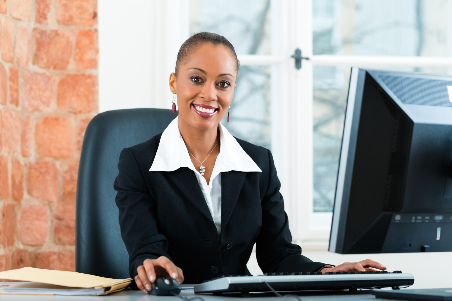 A woman sitting at a desk in an office, smiling at the camera, with a computer monitor, keyboard, and mouse in front of her.