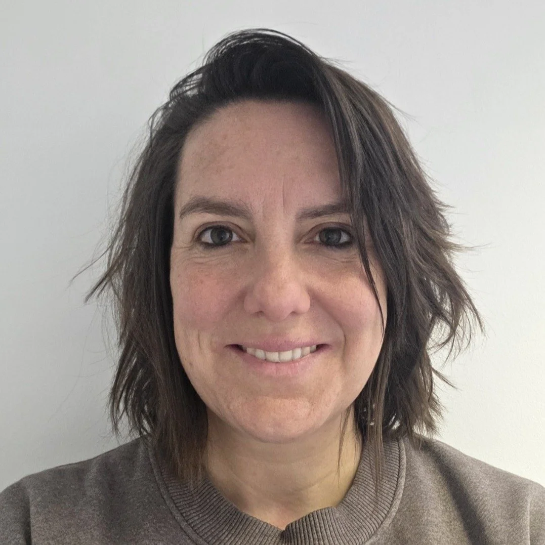 A woman with shoulder-length dark brown hair, blue eyes, and fair skin, smiling at the camera against a plain white background.