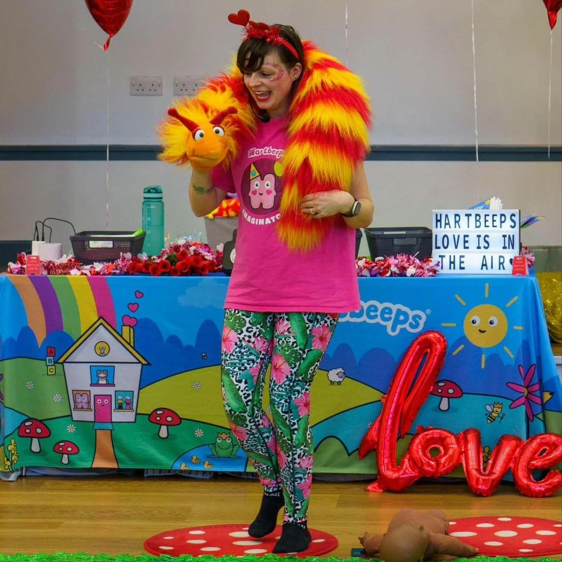 A woman dressed in colorful pajama pants and a pink shirt holding a Kipper puppet. She has a bright orange and yellow feathered scarf, a red headband with a heart, and face paint. Behind her is a decorated table with a sign that reads "HARTBEES LOVE IS IN THE AIR" and various party decorations. The scene appears festive, with balloons and playful colorful decorations.