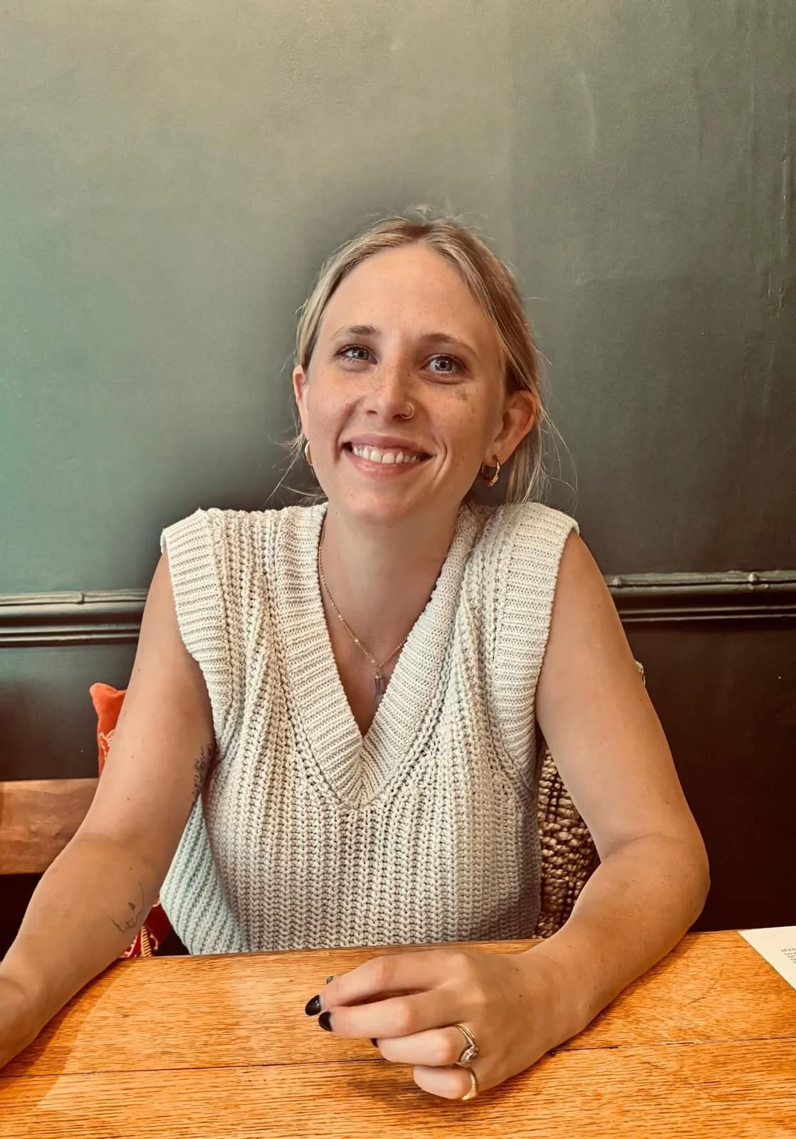 A woman with blonde hair, wearing a knitted beige sleeveless top, sitting at a wooden table in a restaurant, smiling at the camera.
