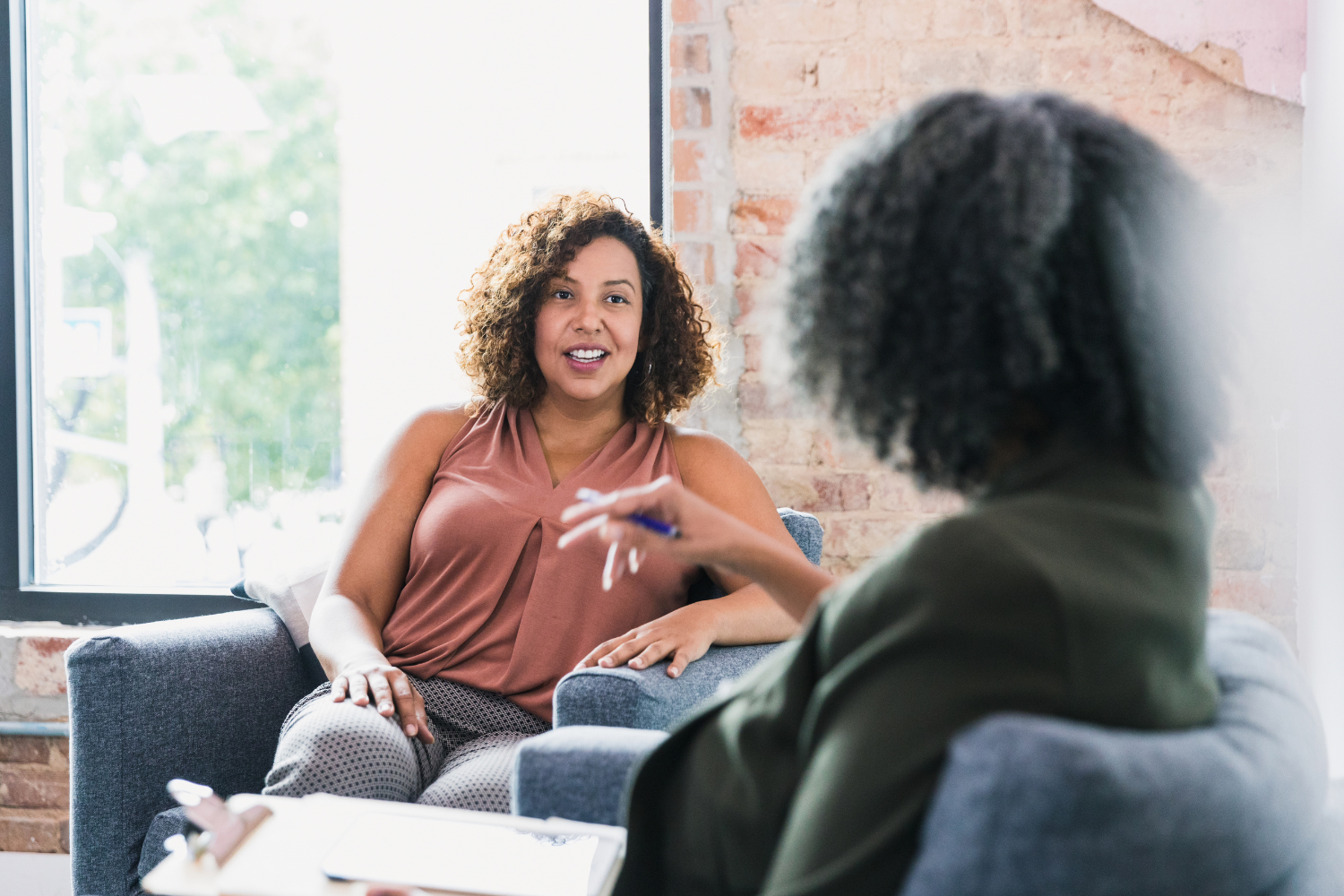 Two women having a conversation in a cozy, well-lit office with brick walls and large window