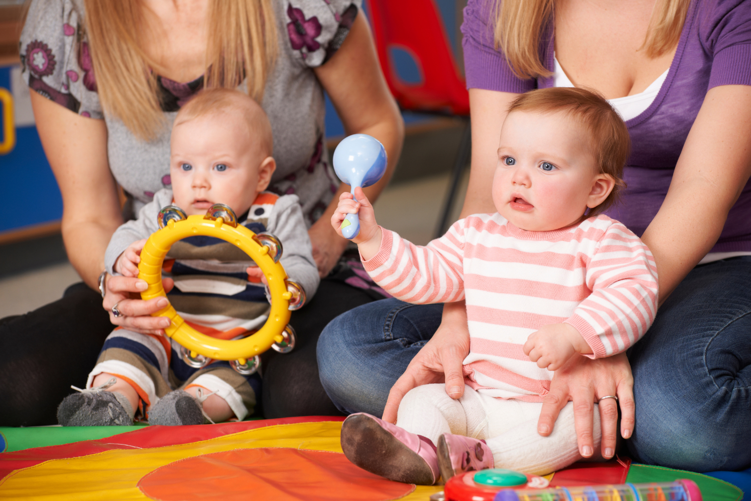 Two babies sitting on a colorful play mat with two women beside them. One baby holds a yellow tambourine, and the other holds a blue rattle, with toys scattered on the mat.