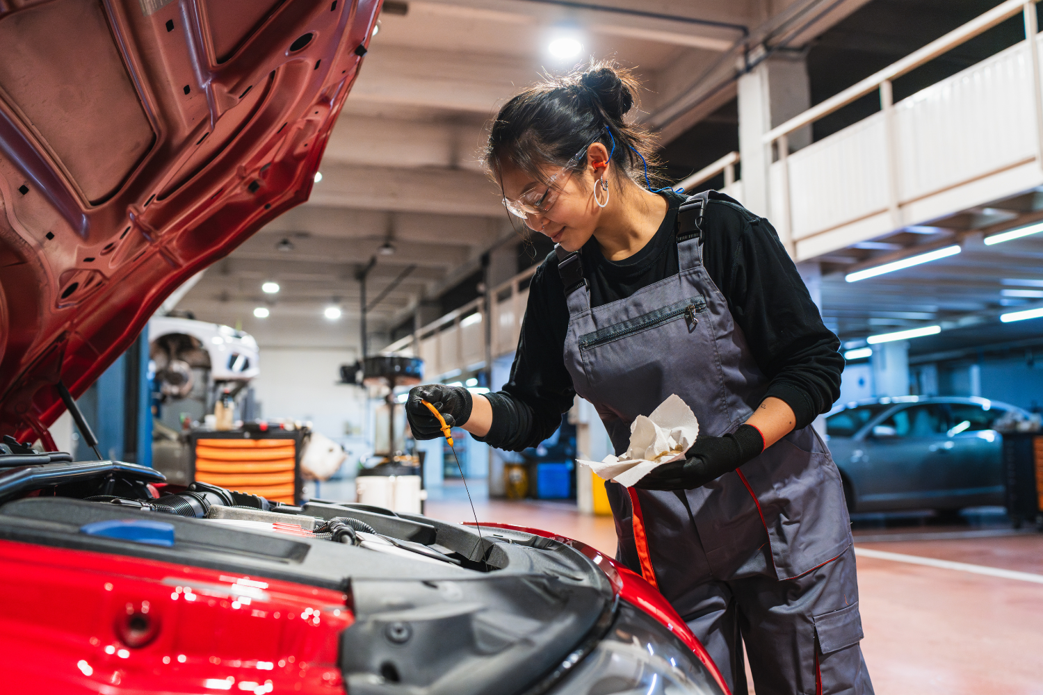 A female mechanic wearing safety glasses, ear protection, and overalls working on a red car engine inside an auto repair shop.