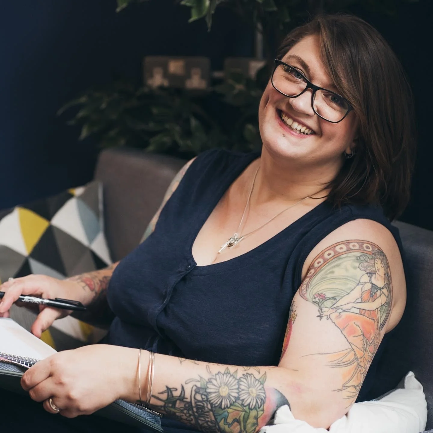 A woman with glasses smiling, showing tattoos on her arm and shoulder, sitting indoors with a dark background and patterned cushion.