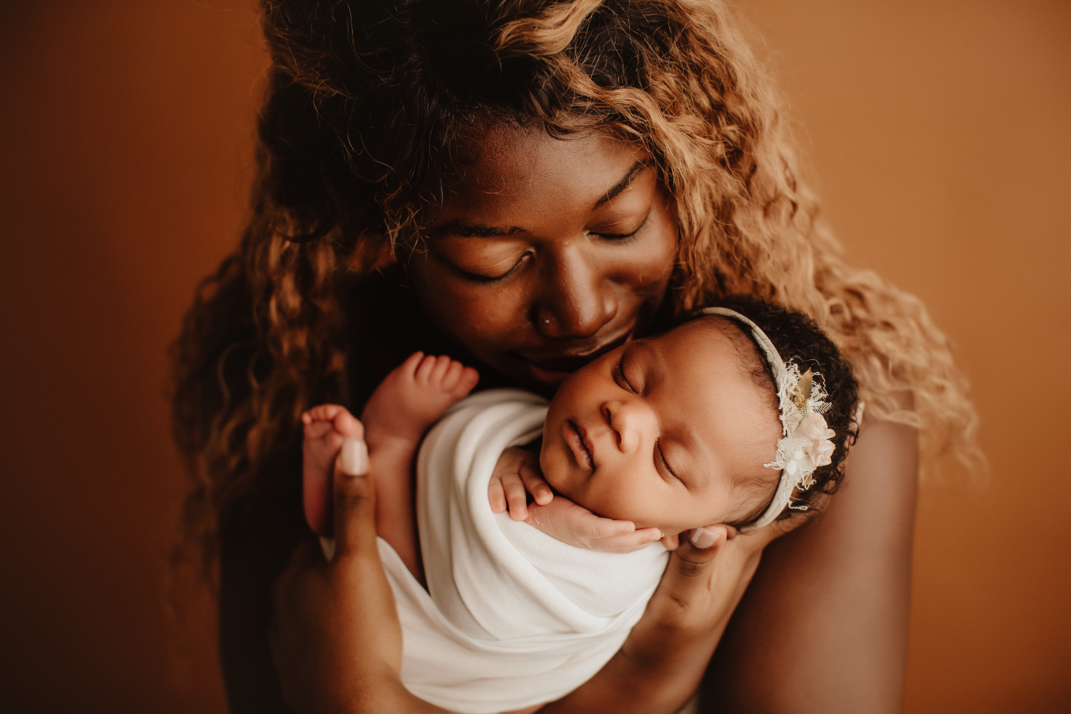 A woman with curly hair holding a sleeping baby close to her face against a warm brown background.