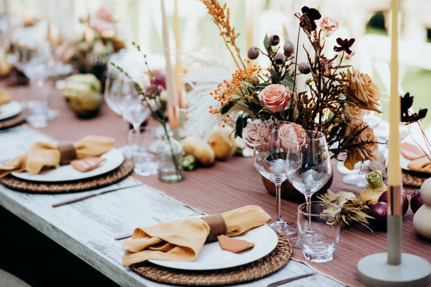 A table set for a special occasion with plates, napkins, glasses, and a floral centerpiece featuring pink roses, dried flowers, and berries, with tall yellow candles and decorative items.