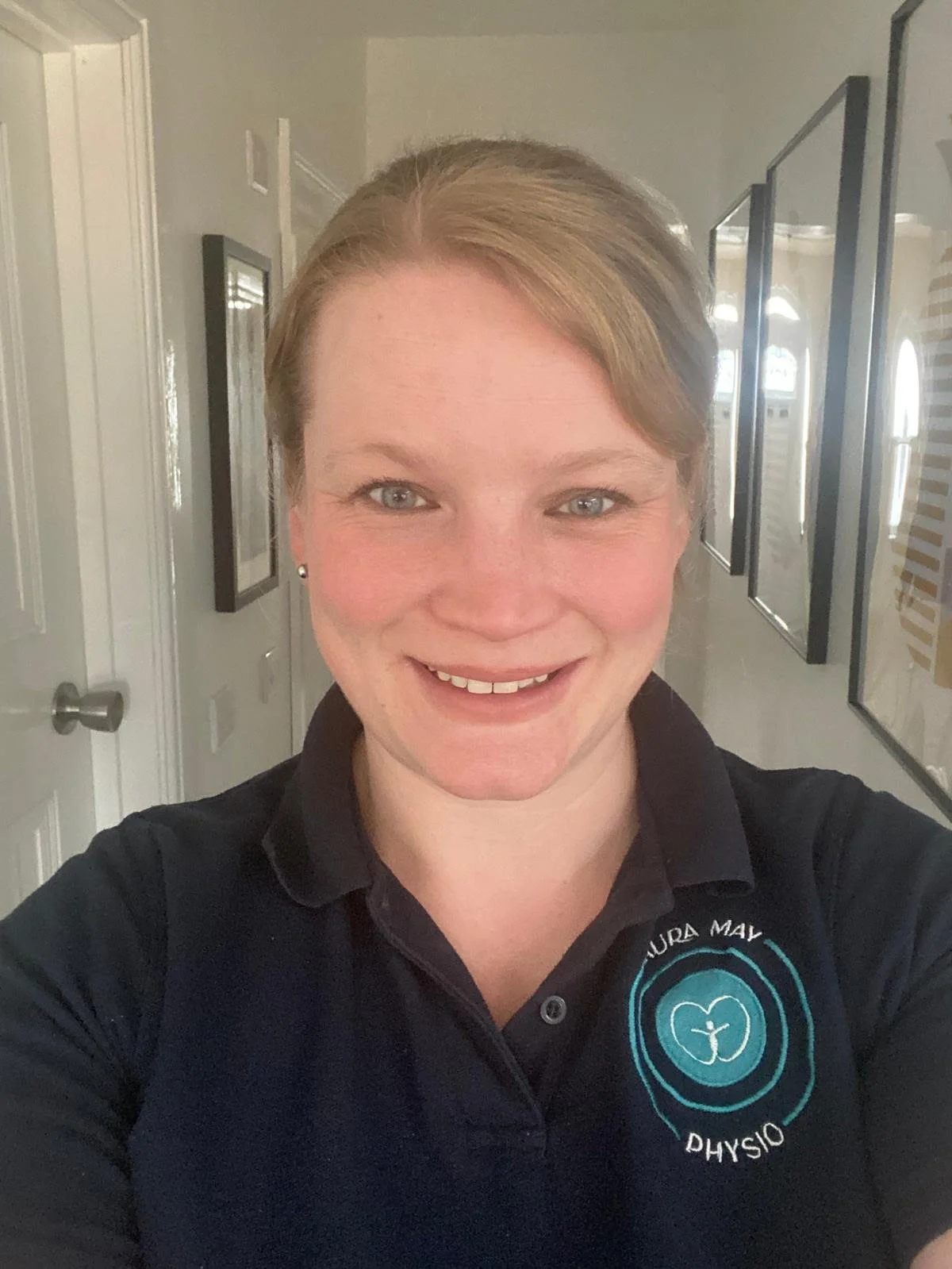 A woman taking a selfie in a hallway, smiling, wearing a black polo shirt with a logo that says 'Laura May Physical Therapy.'