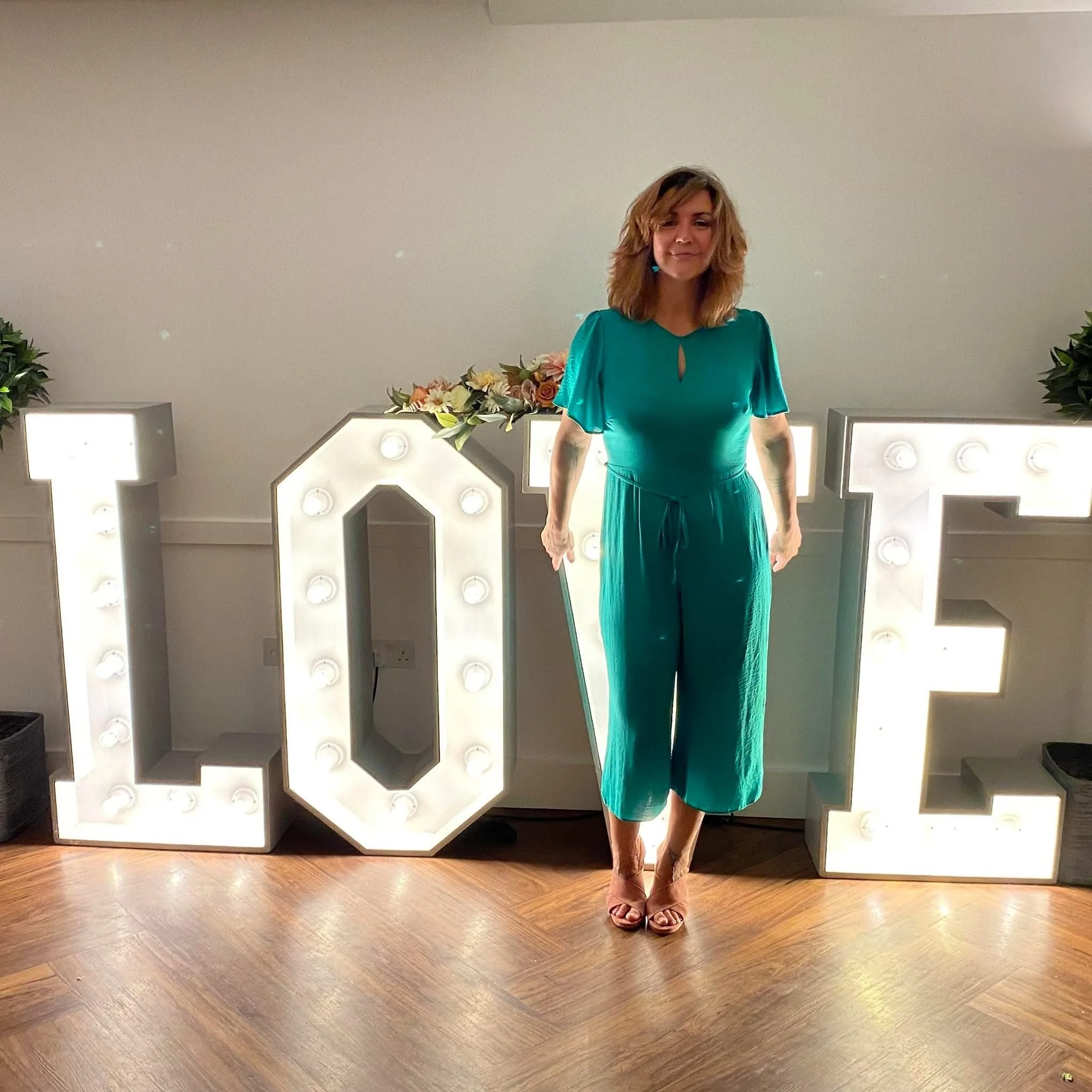 A woman in a teal dress standing in front of backlit illuminated letters with flowers on top and potted plants on either side.