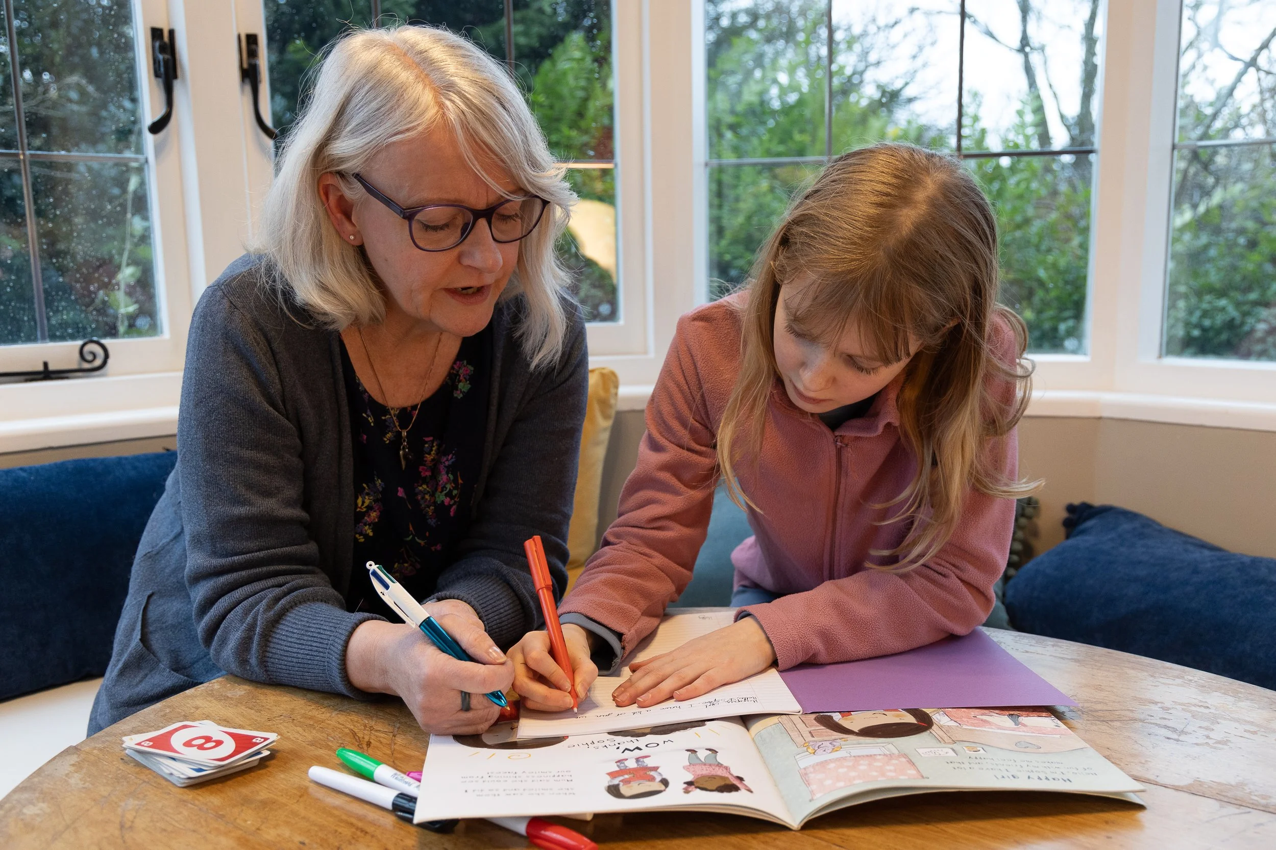 An older woman and a young girl are sitting at a wooden table, looking at an open book and writing or drawing in it with markers. The woman has gray hair, glasses, and is wearing a gray cardigan, while the girl has long, blonde hair and is wearing a pink sweatshirt. There are some coloring books, markers, and cards on the table. They are in a room with large windows showing trees outside.