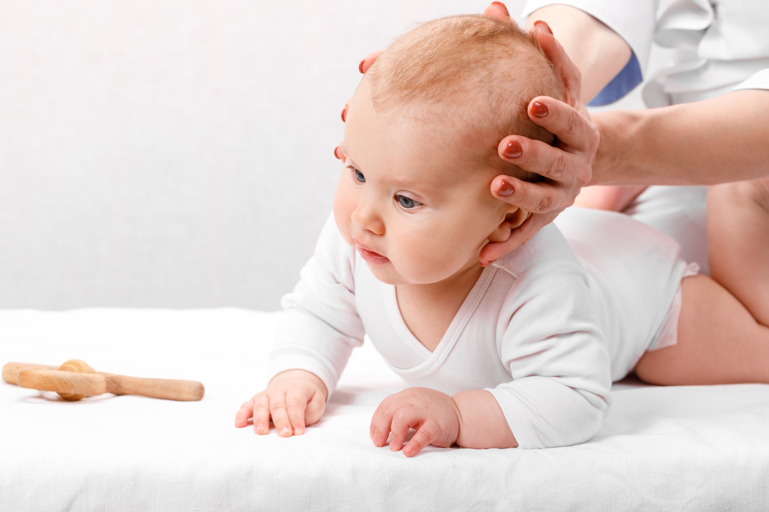 A baby is lying on their stomach on a white bed, being examined by a healthcare professional. The healthcare worker gently holds the baby's head and neck. A wooden rattle toy is on the bed nearby.