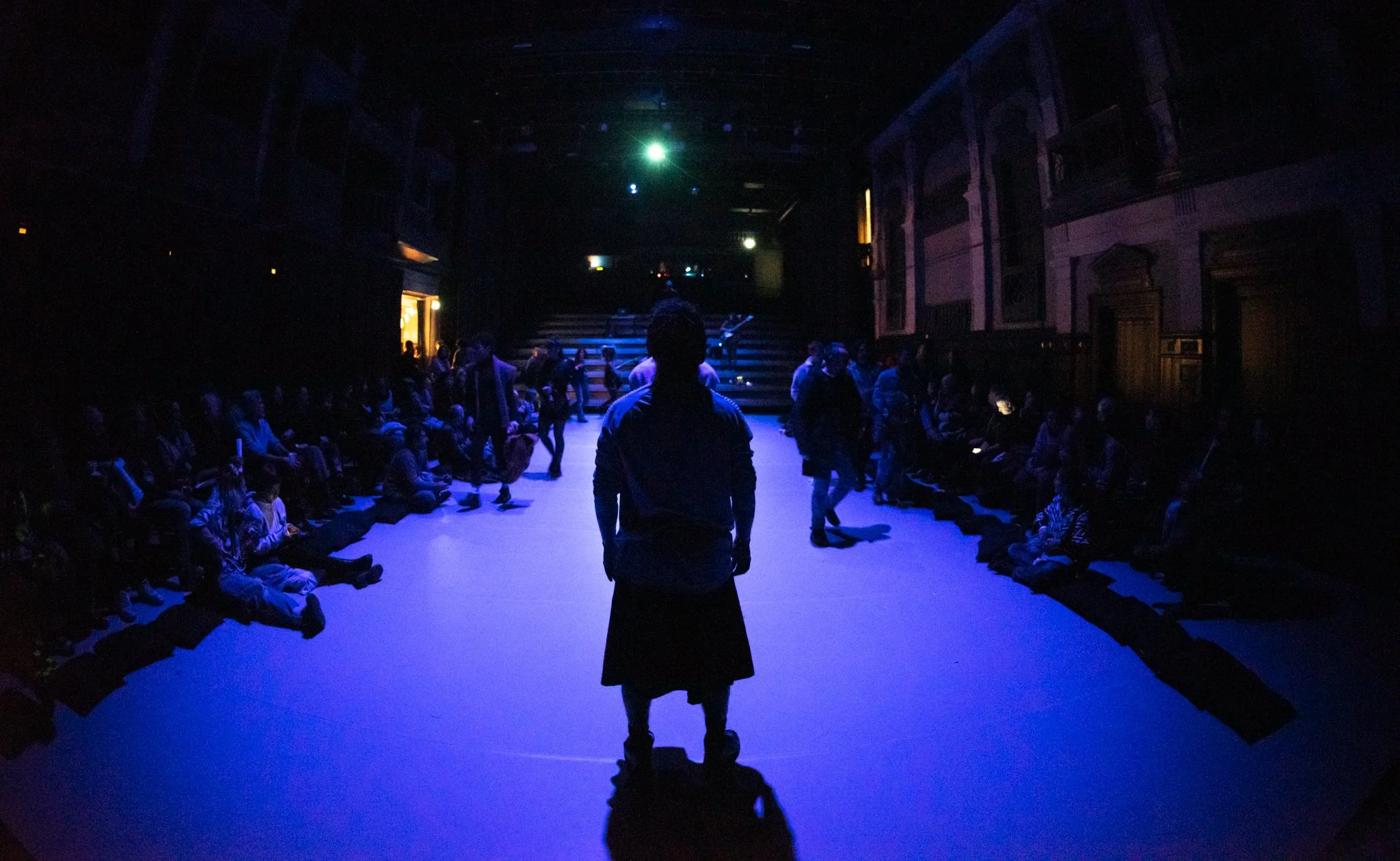 Dark theater with a small stage, audience seated on floor and steps, a figure in front of the stage with their back to the camera, brightly colored lights overhead.