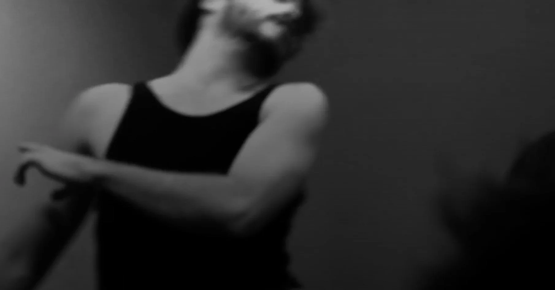 A black and white photo of a woman with crossed arms, wearing a sleeveless top, standing against a dark background.