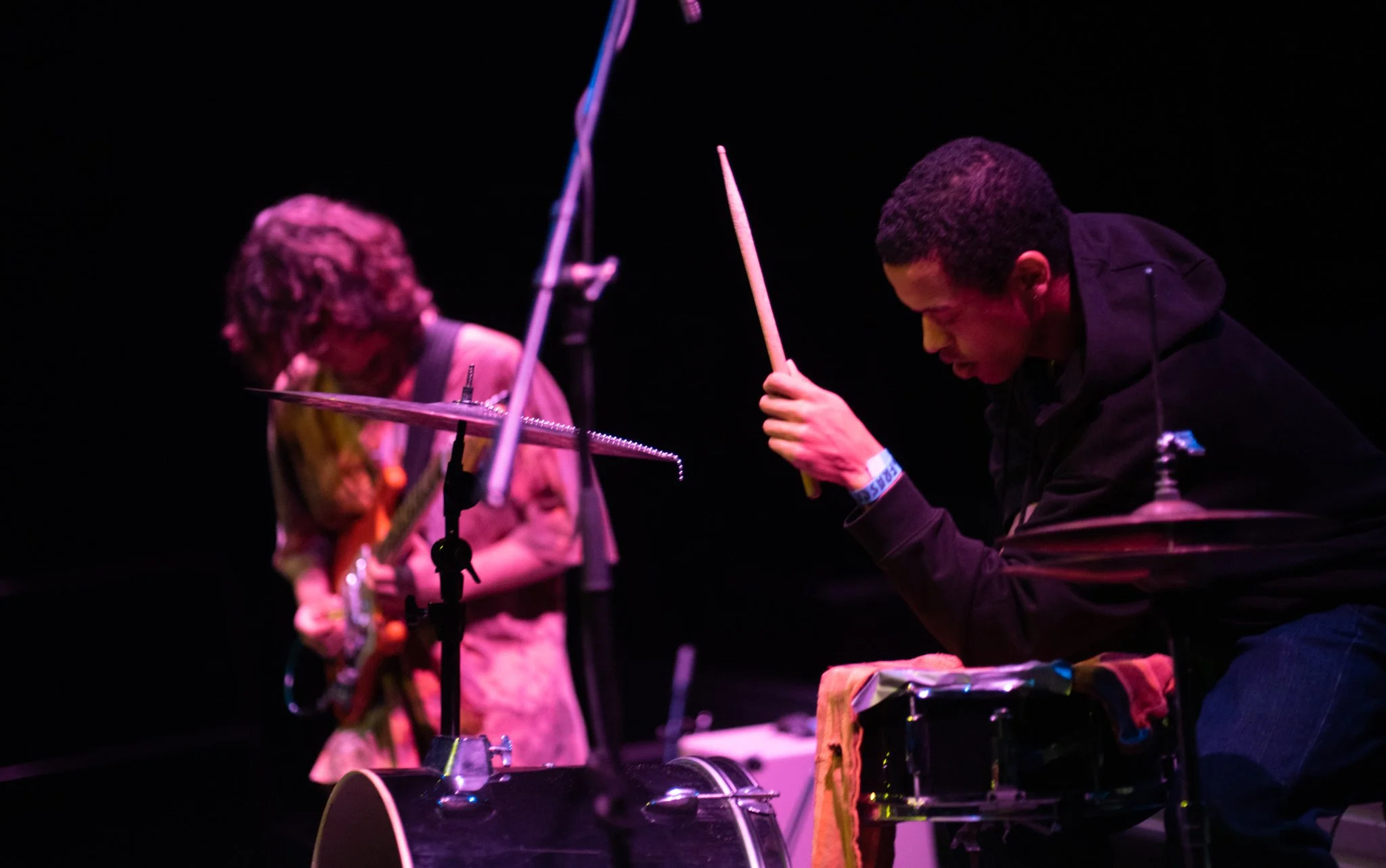 Two musicians performing on stage with dark background, one playing guitar and the other playing drums, under pink stage lighting.