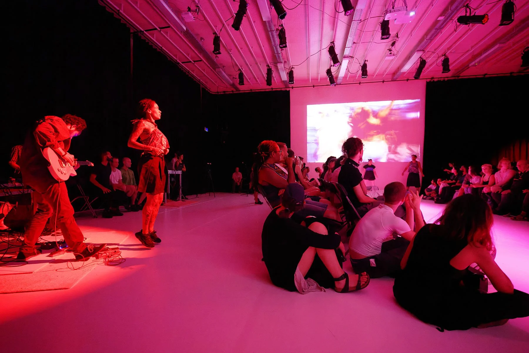 An indoor stage with pink lighting shows musicians performing, with an audience seated on the floor and in chairs, watching attentively.
