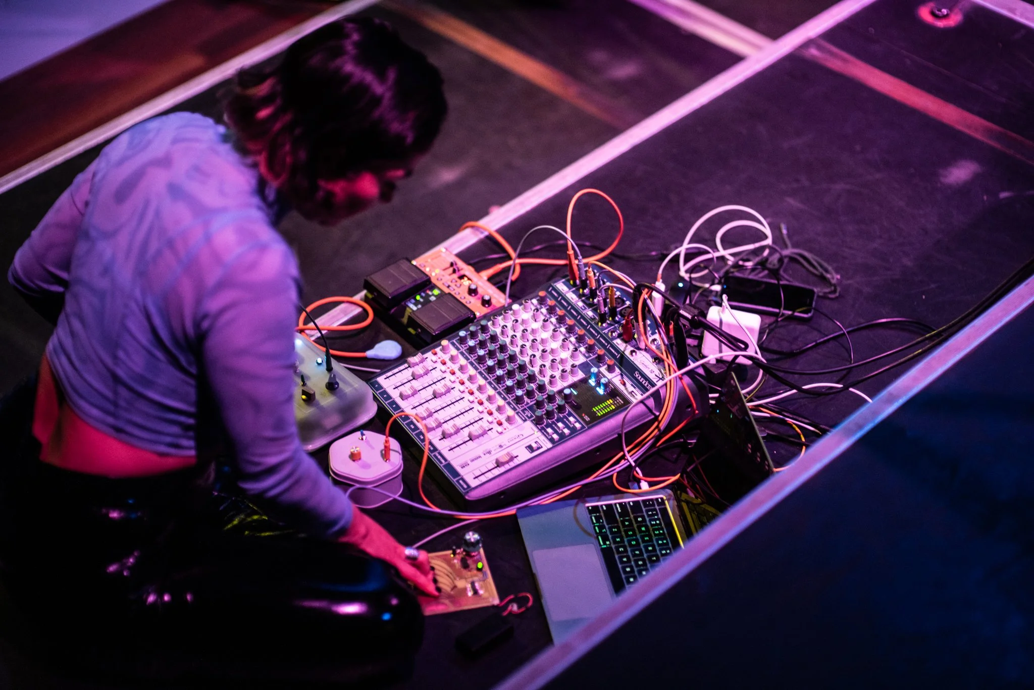 A person with dark hair playing electronic music using a mixer, laptop, and effects pedals in a dimly lit venue with purple and pink lighting.