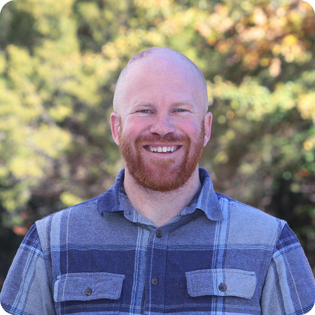 A smiling man with a shaved head and red beard standing outdoors with blurred trees and foliage in the background.