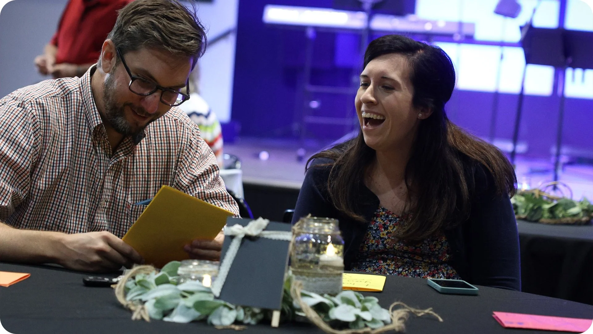 A man and woman sitting at a decorated table, smiling and laughing; the man is looking at a yellow piece of paper, and the woman is looking at him. The table has a candle in a jar, a small makeup mirror, and colorful notes.