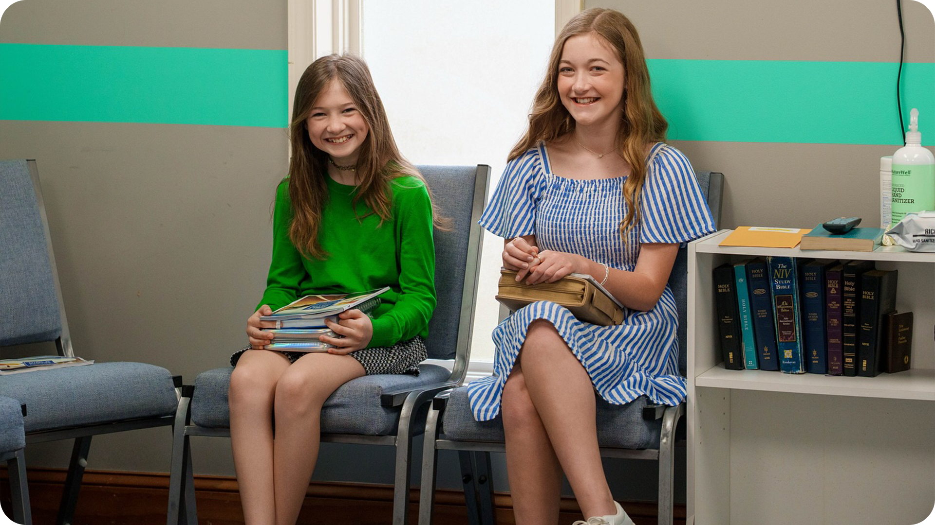 Two young girls sitting on chairs in a waiting room, smiling and holding magazines or books, with bookshelves and sanitizer on a side table behind them.