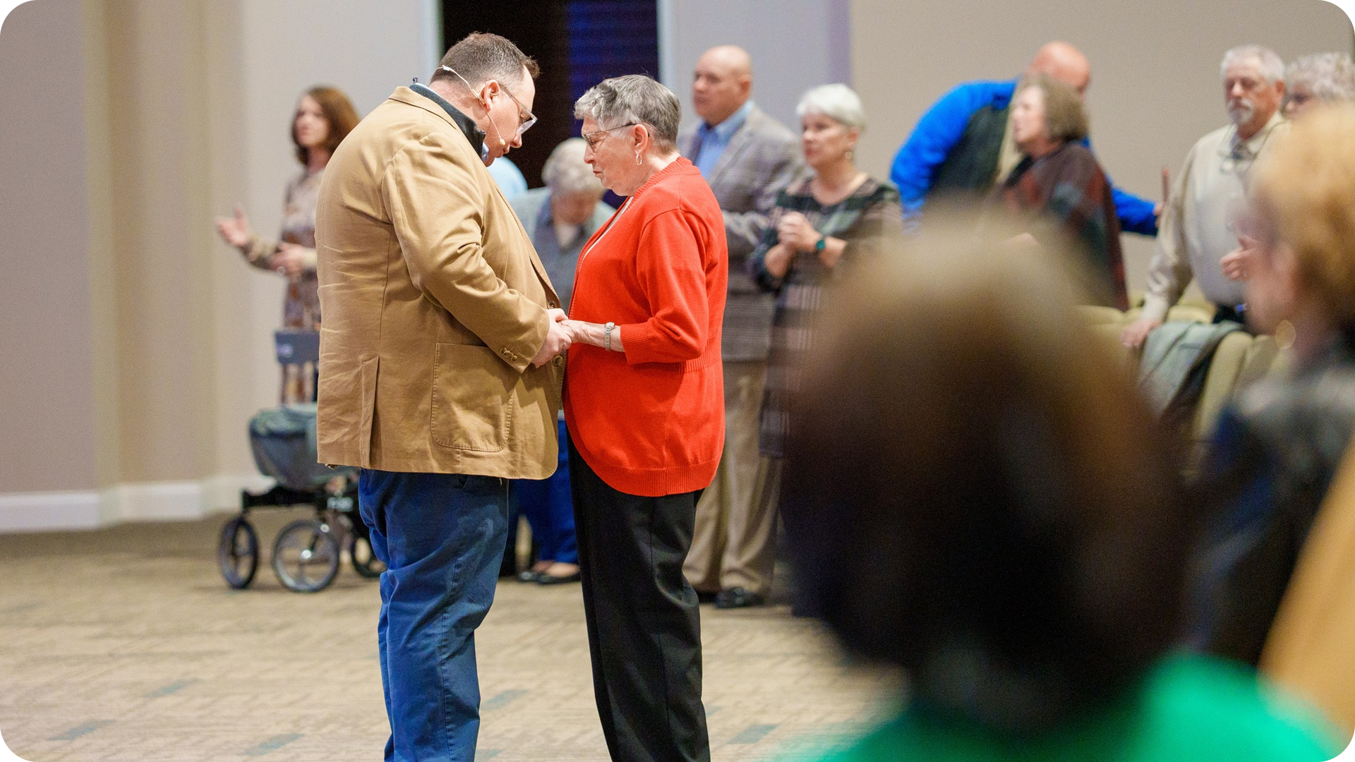 A man and woman hold hands and pray surrounded by people standing and sitting in a room.