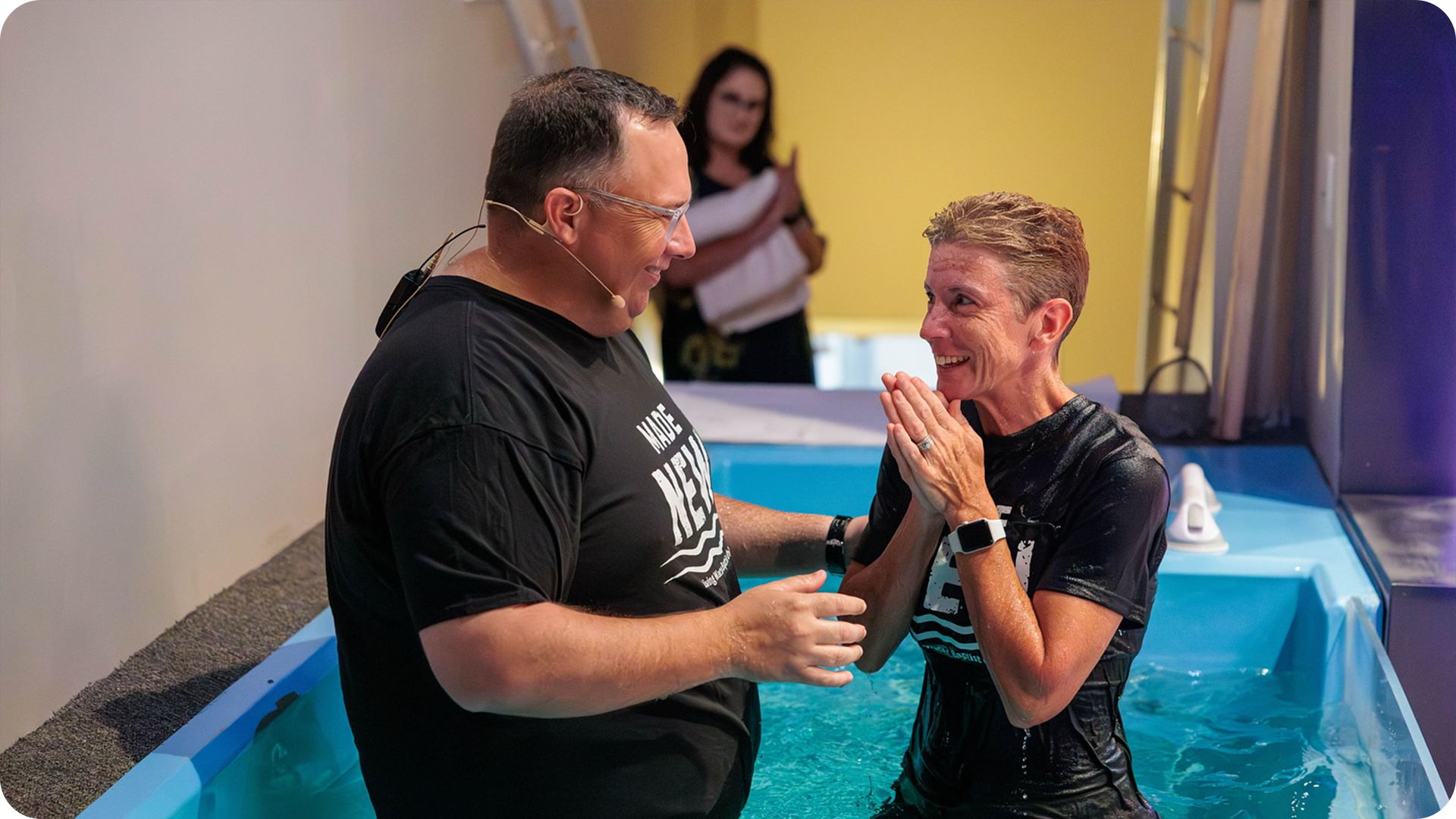 A man and woman in a water baptism pool, smiling and holding hands as the man baptizes the woman. A third person is in the background, clapping.