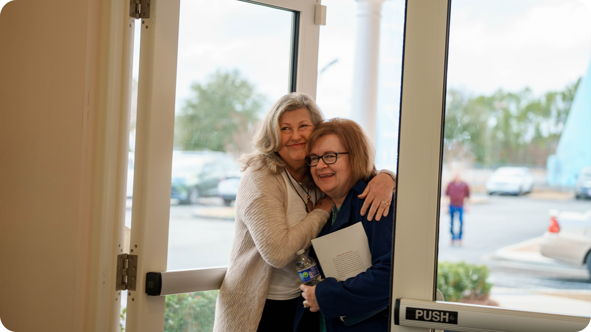Two women hugging at the door, smiling, in a cheerful scene outside a building.