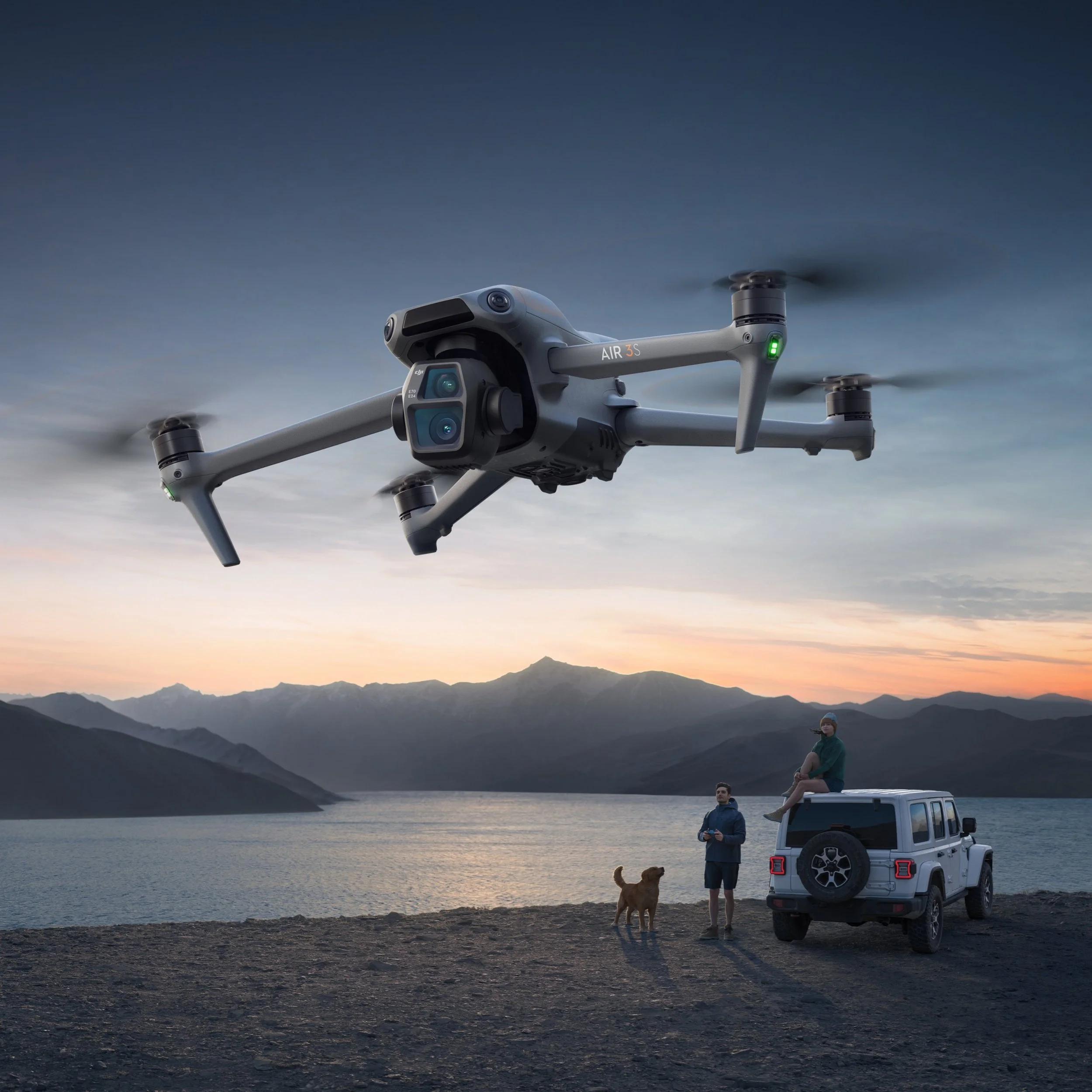 A DJI Air 3S drone flying in the sky over a scenic landscape with mountains and a lake during sunset, with two people and a dog standing near a white Jeep vehicle near the shore.