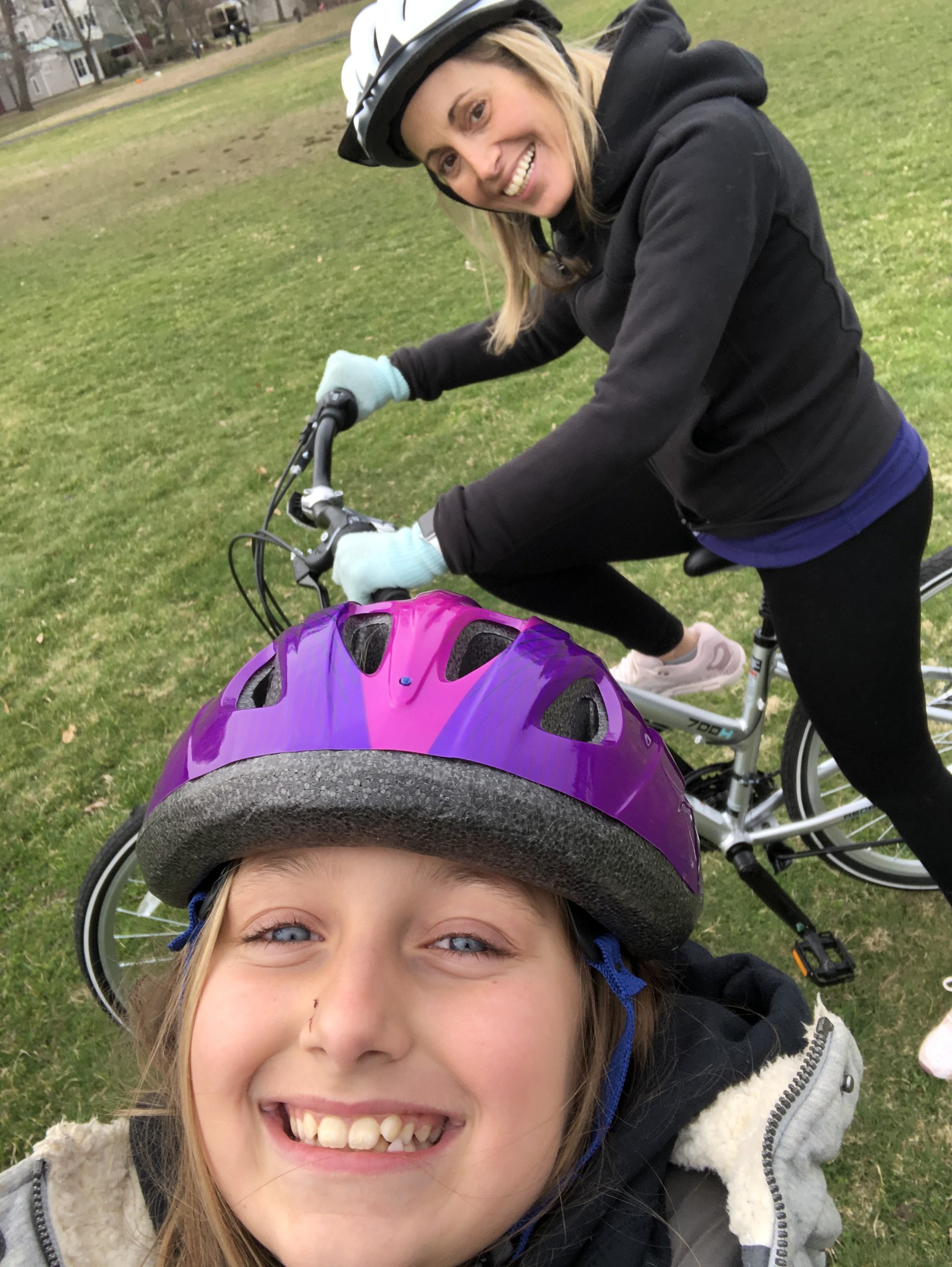 A smiling young girl in a helmet taking a selfie with two women in the background, also wearing helmets, riding bikes on a grassy field with houses in the distance.