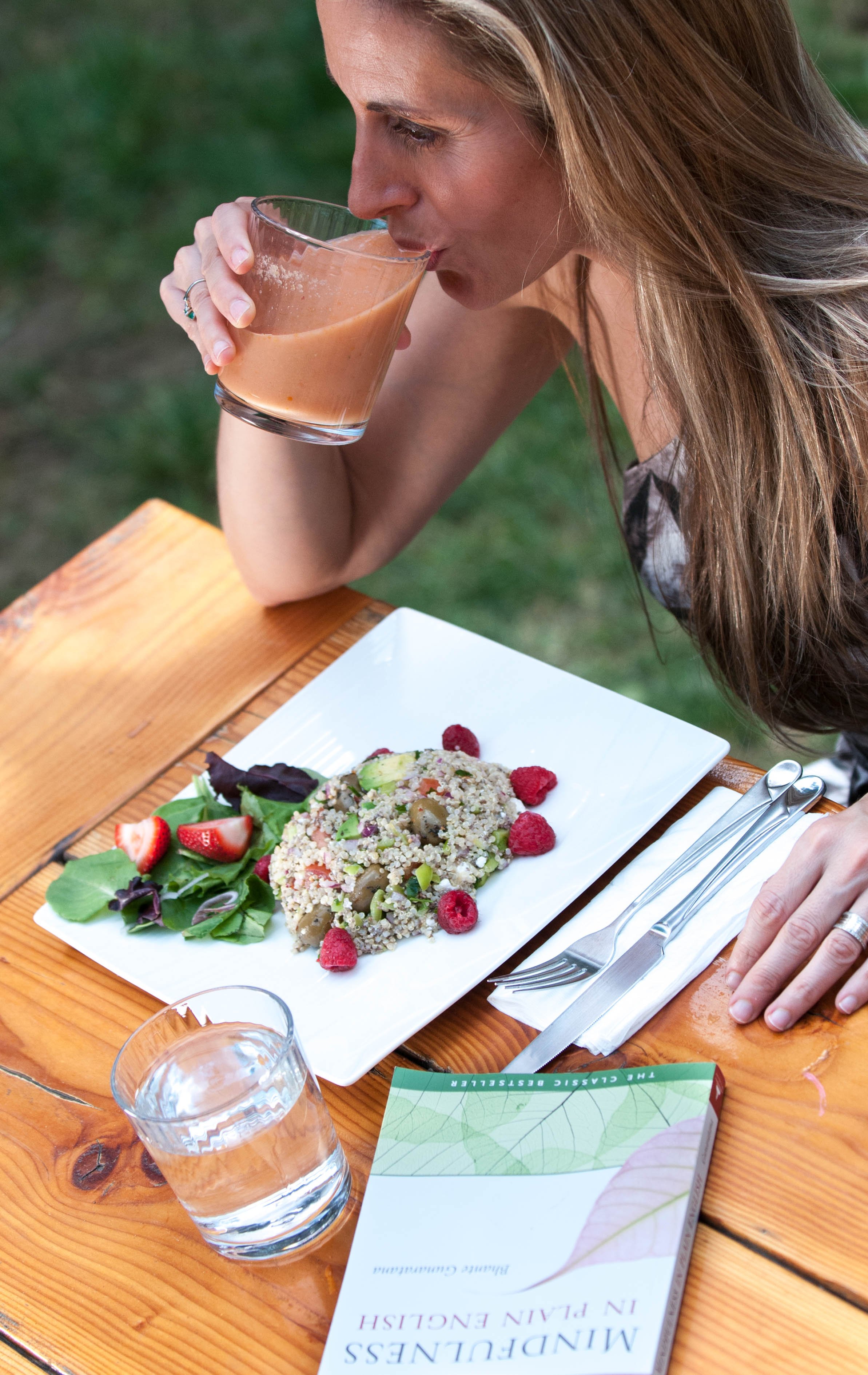 A woman with long light brown hair is drinking a pink smoothie from a glass. There is a rectangular white plate with a grain salad garnished with raspberries and strawberries, and a side of greens and sliced strawberries on the wooden table. A glass of water and a book titled "Mindfulness in Plant-Based Eating" are also on the table.