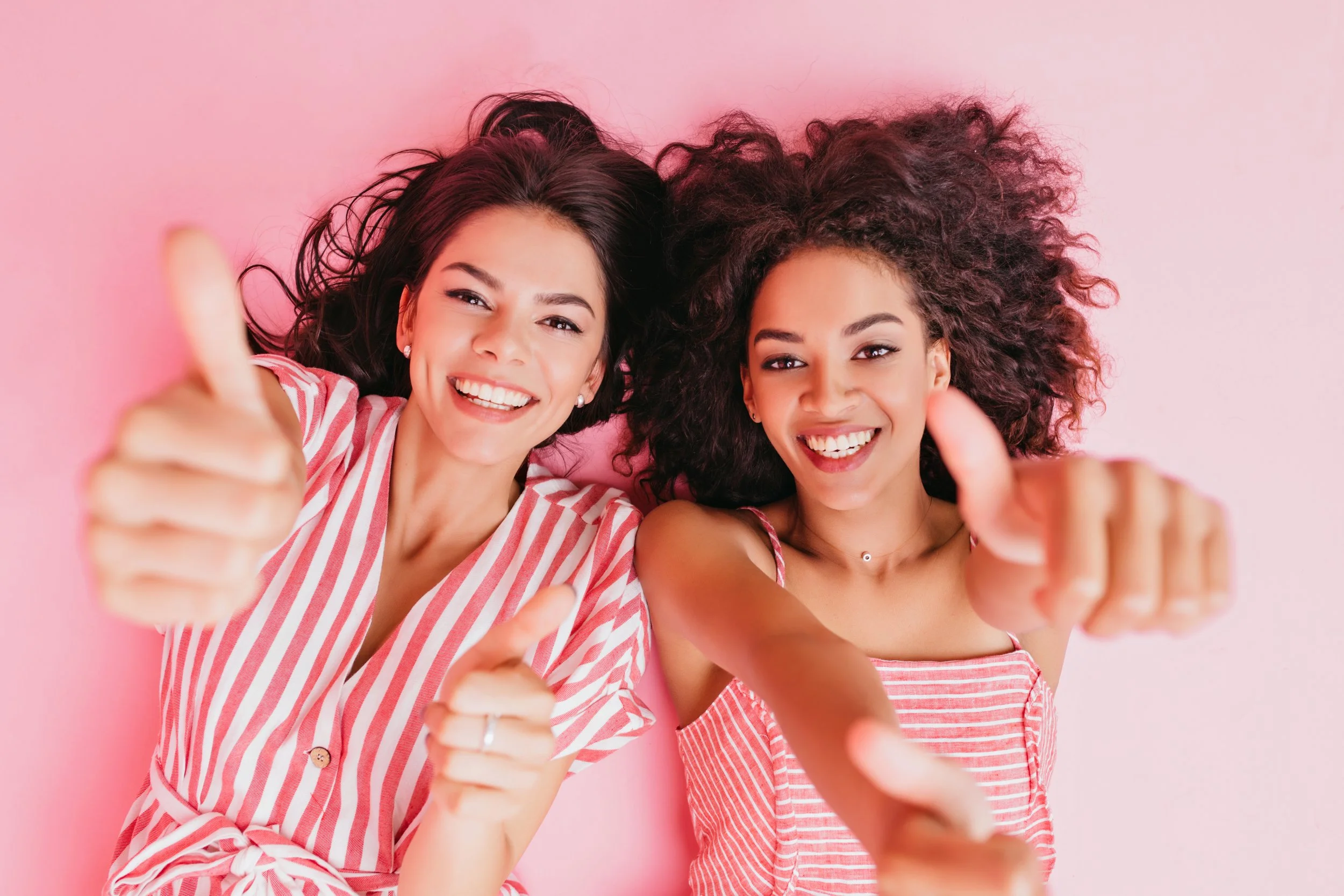 Two women lying on a pink background taking a selfie, smiling, and giving thumbs up.