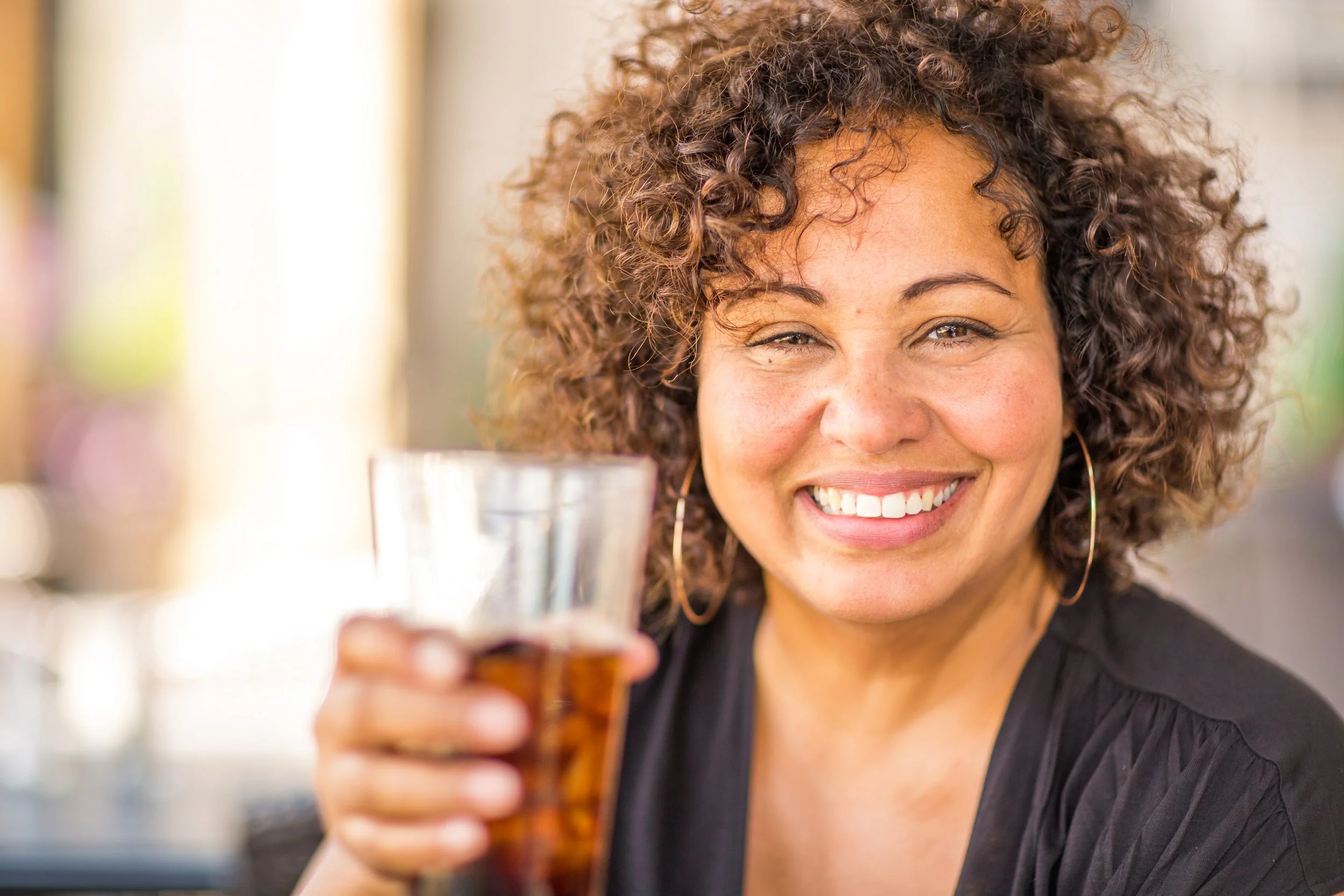 Smiling woman with curly hair holding a glass of dark soda, wearing hoop earrings and a black top.