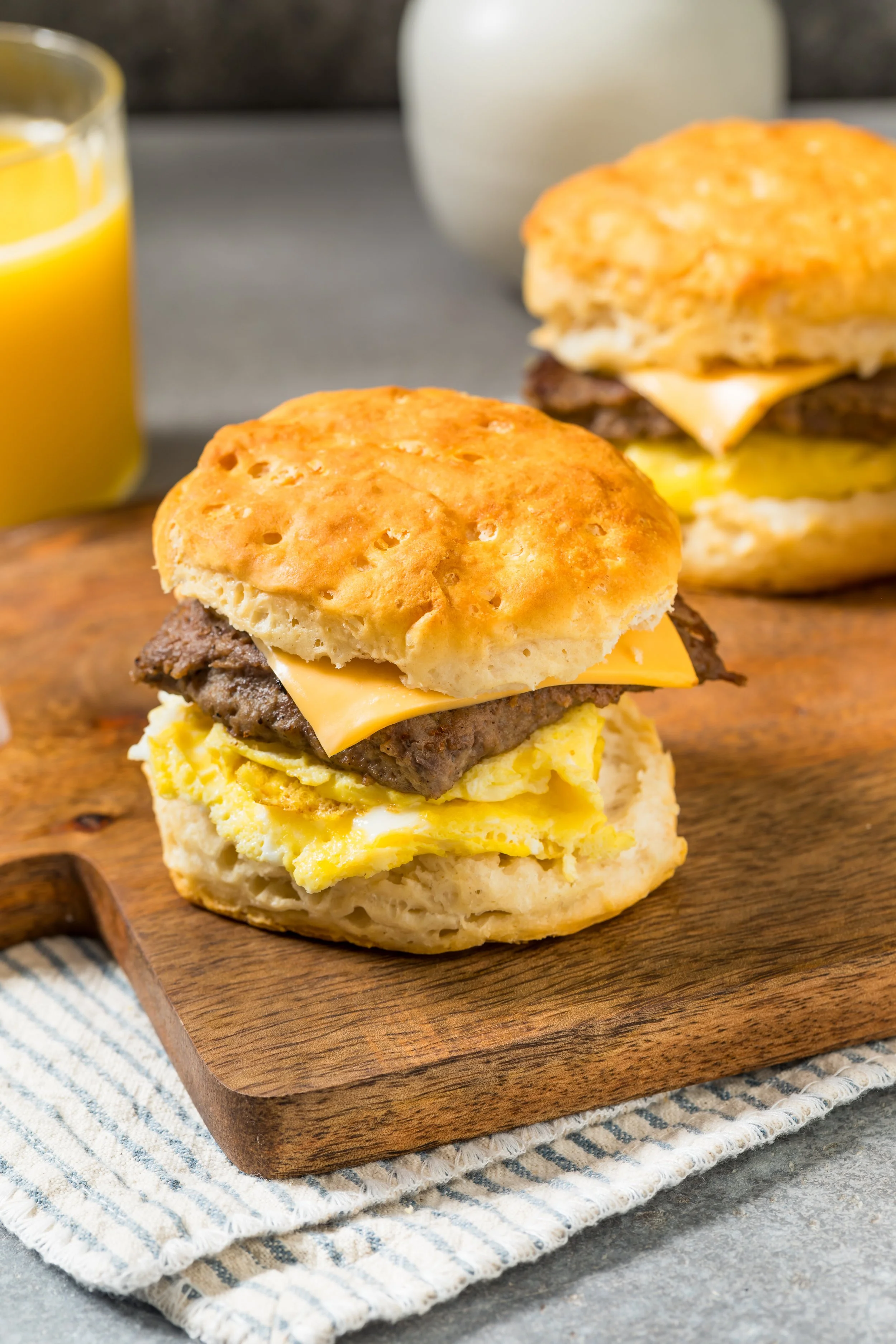 Close-up of a breakfast sandwich with scrambled eggs, cheese, sausage patty, and biscuit on a wooden serving board, with a glass of orange juice in the background.