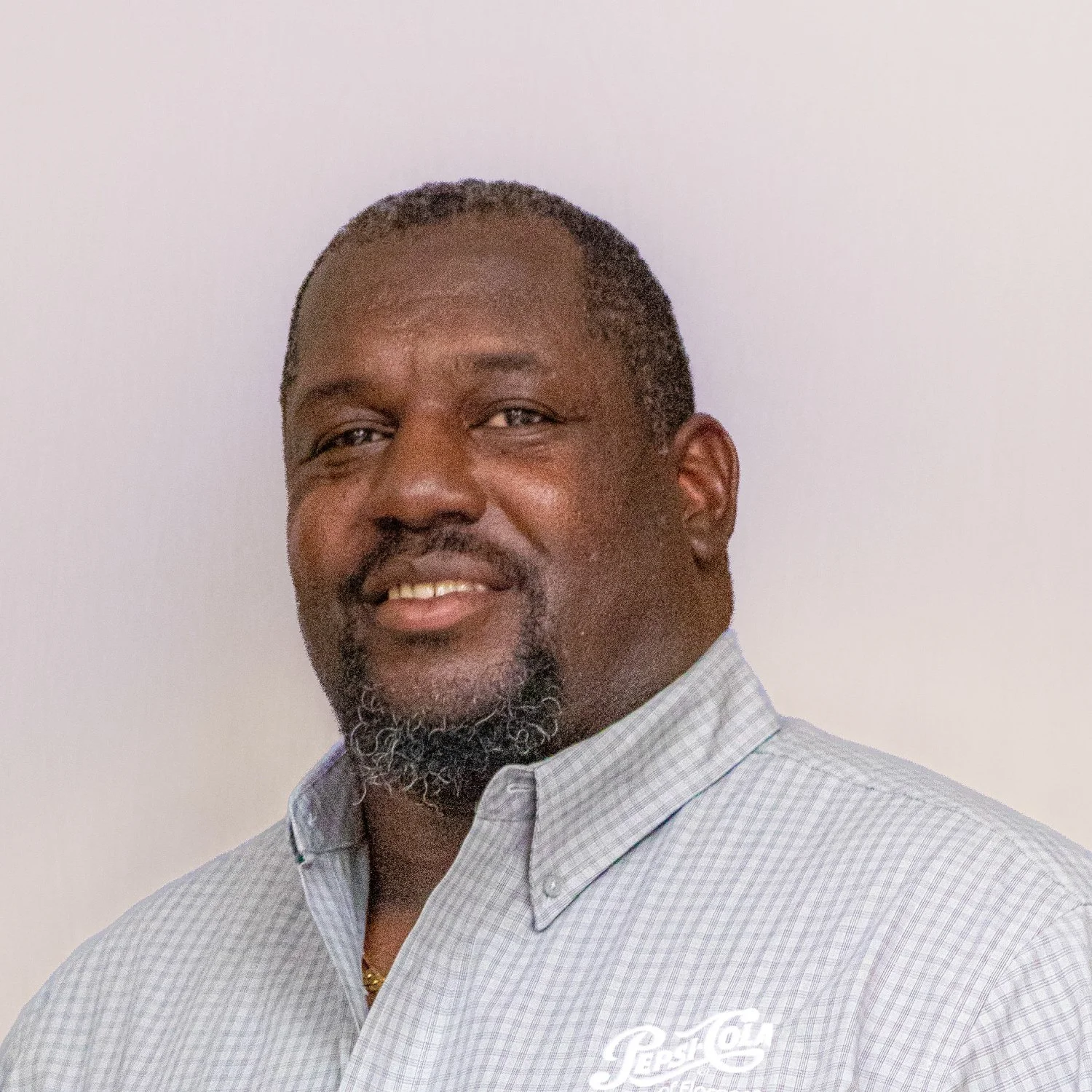 A middle-aged man with dark skin, short hair, and a beard, smiling and wearing a light blue, checkered collared shirt with a Pepsi logo.
