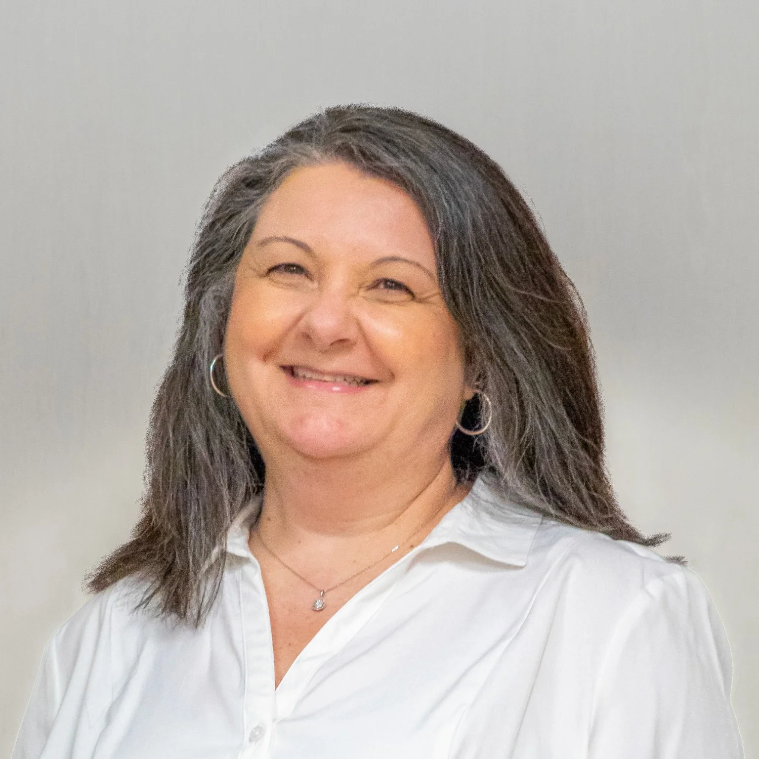 Smiling woman with shoulder-length dark hair, wearing a white shirt, hoop earrings, and a necklace, standing against a plain background.