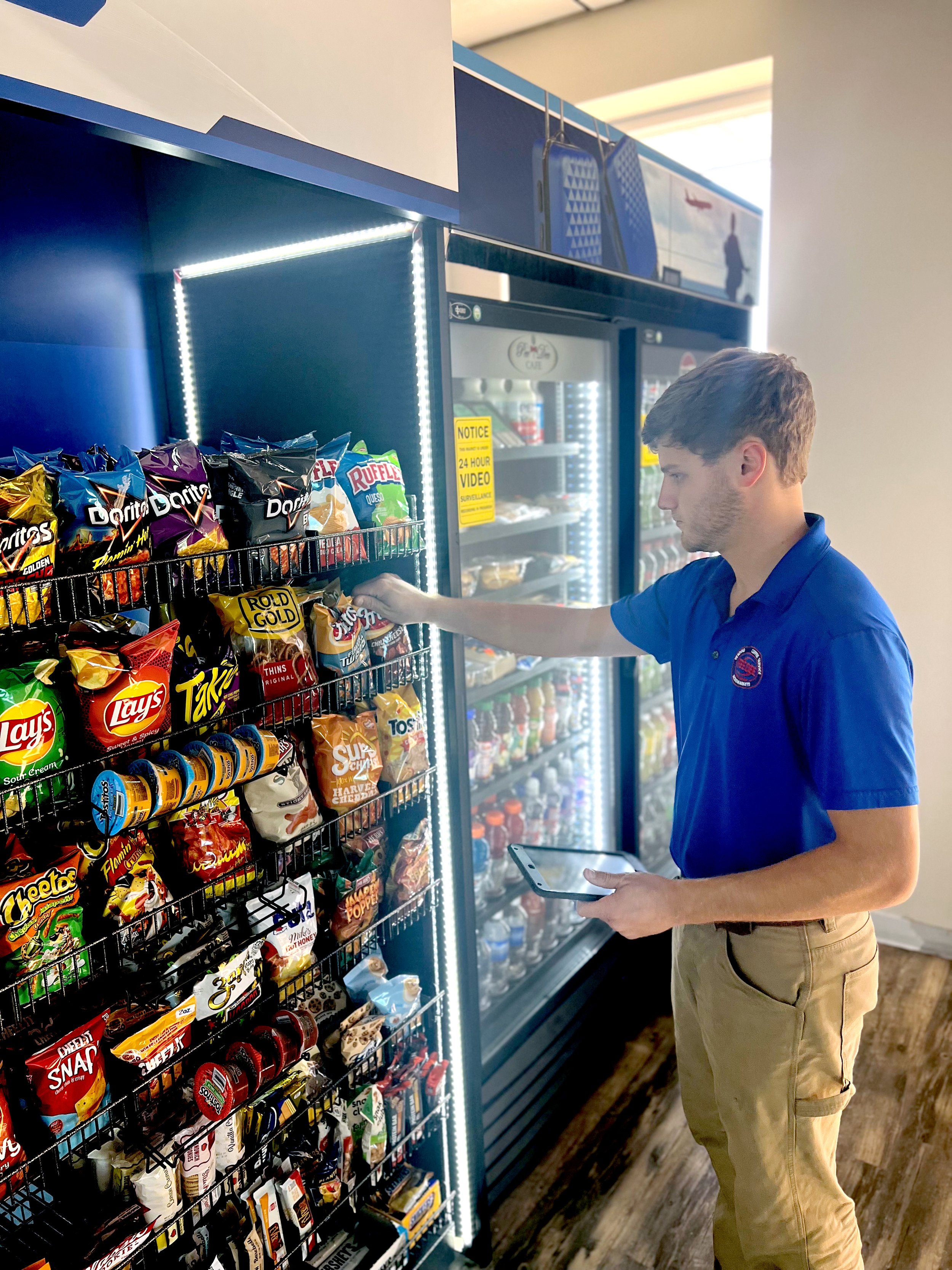 Person in blue polo shirt shopping for snacks in front of vending machines in a convenience store.